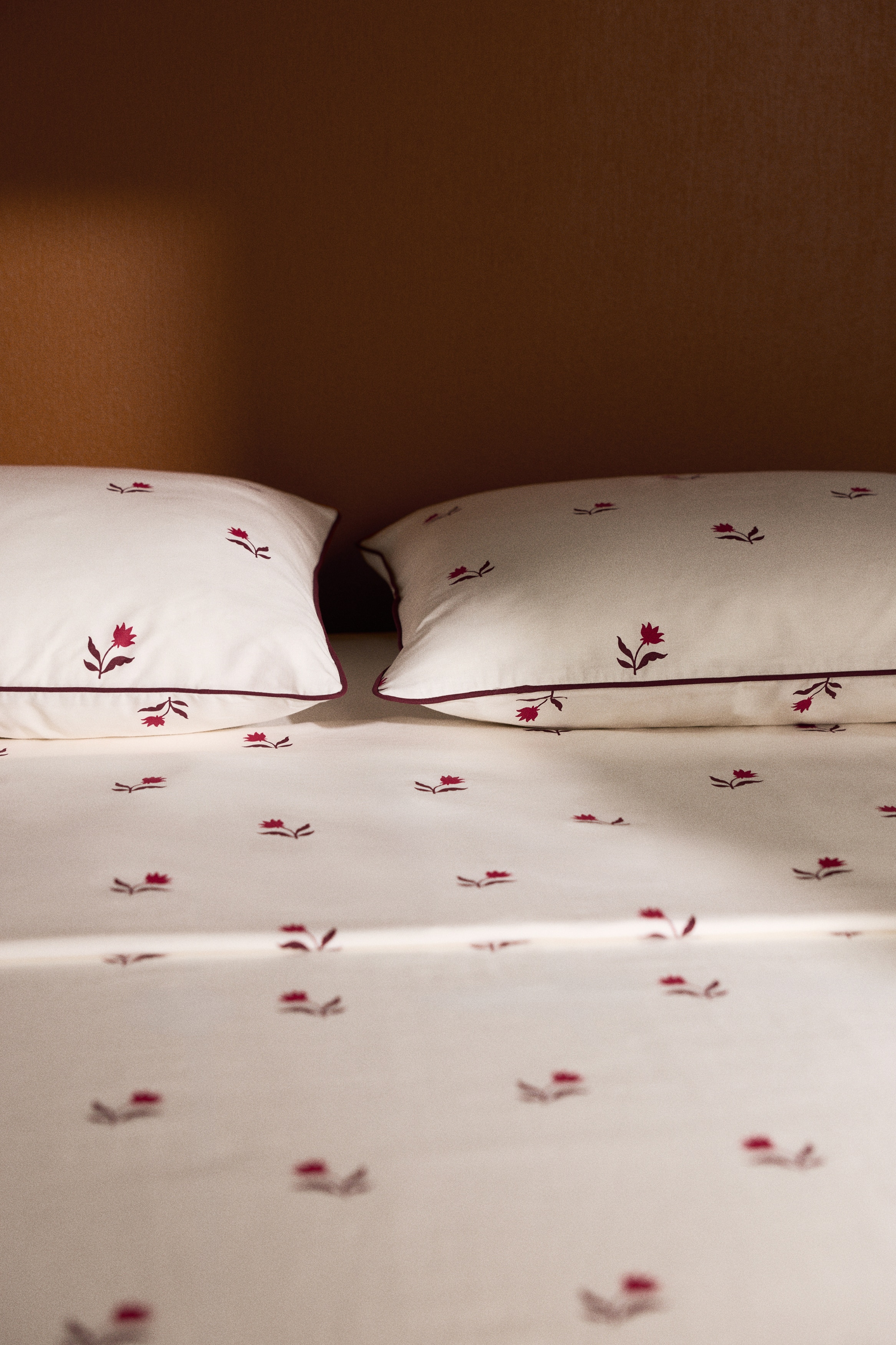 A white bedding set showcases a repeating pattern of small, dark red floral motifs across the duvet cover and the two matching pillowcases. The pillowcases feature thin, dark red piped borders along their edges.