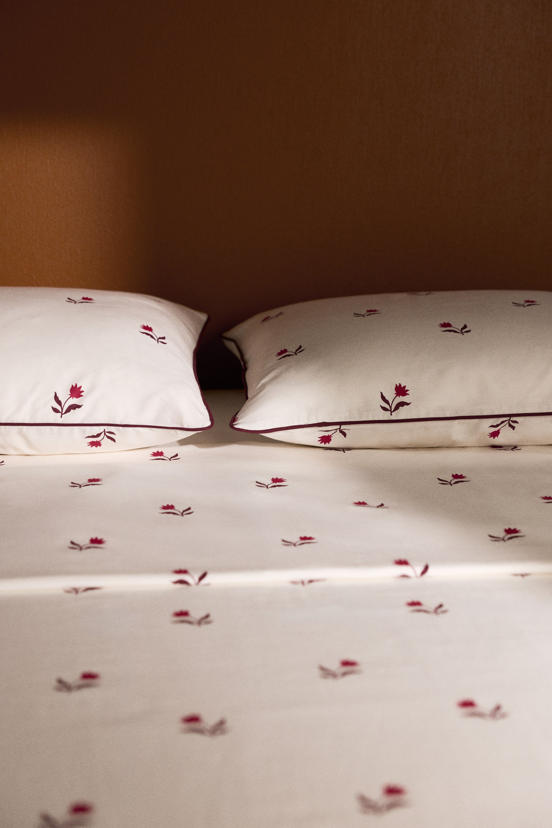 View larger image: A cotton bed sheet set is displaying an off-white flat sheet and two matching pillowcases, all featuring an all-over red floral print. The pillowcases are lying flat side by side, showcasing contrasting dark red piping along their stitched edges.