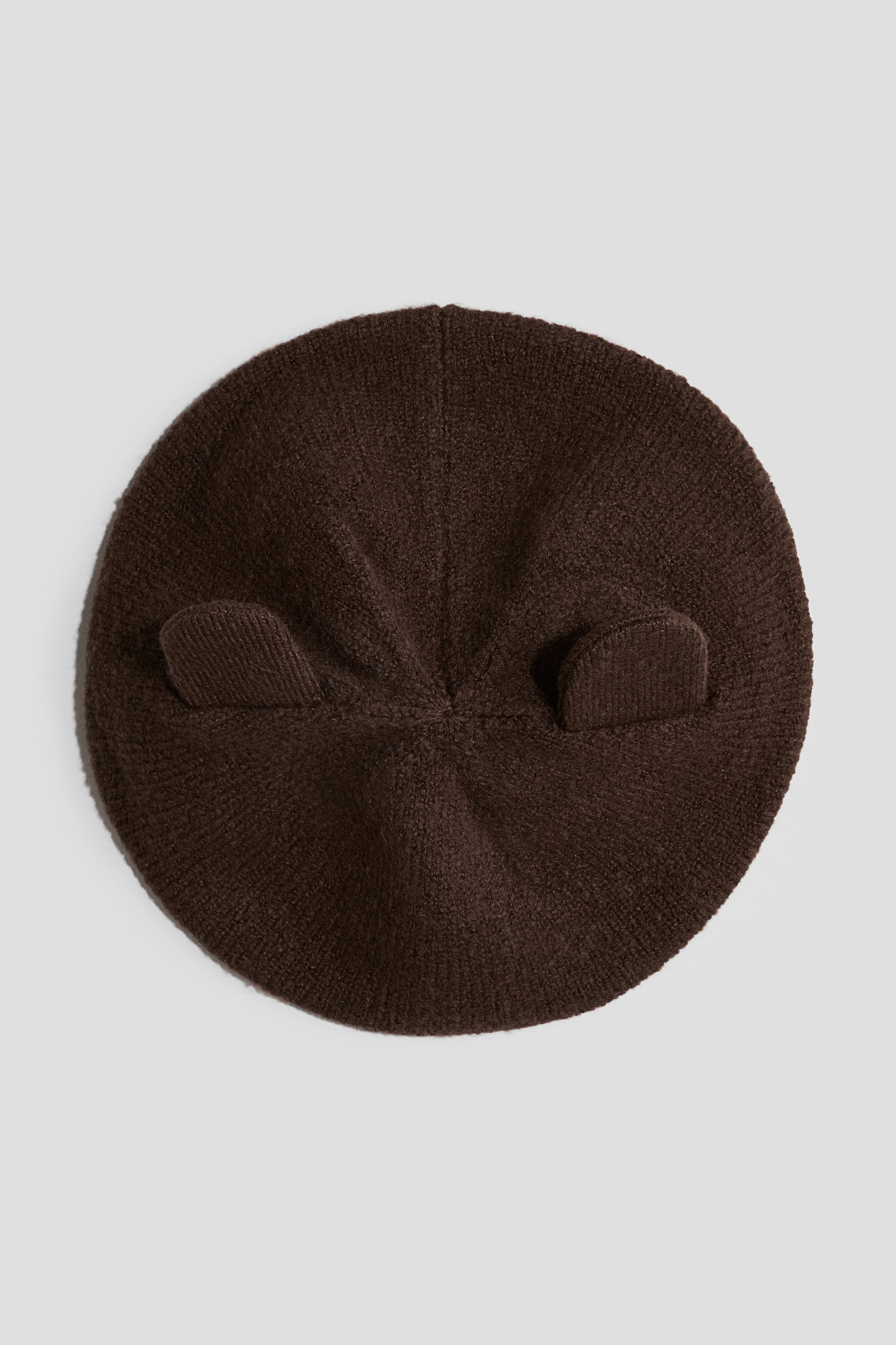 The brown knitted beret is being worn tilted on a modelâs head, showcasing a soft, textured knitted construction with a ribbed brim. It features a small decorative knitted stalk on the top.