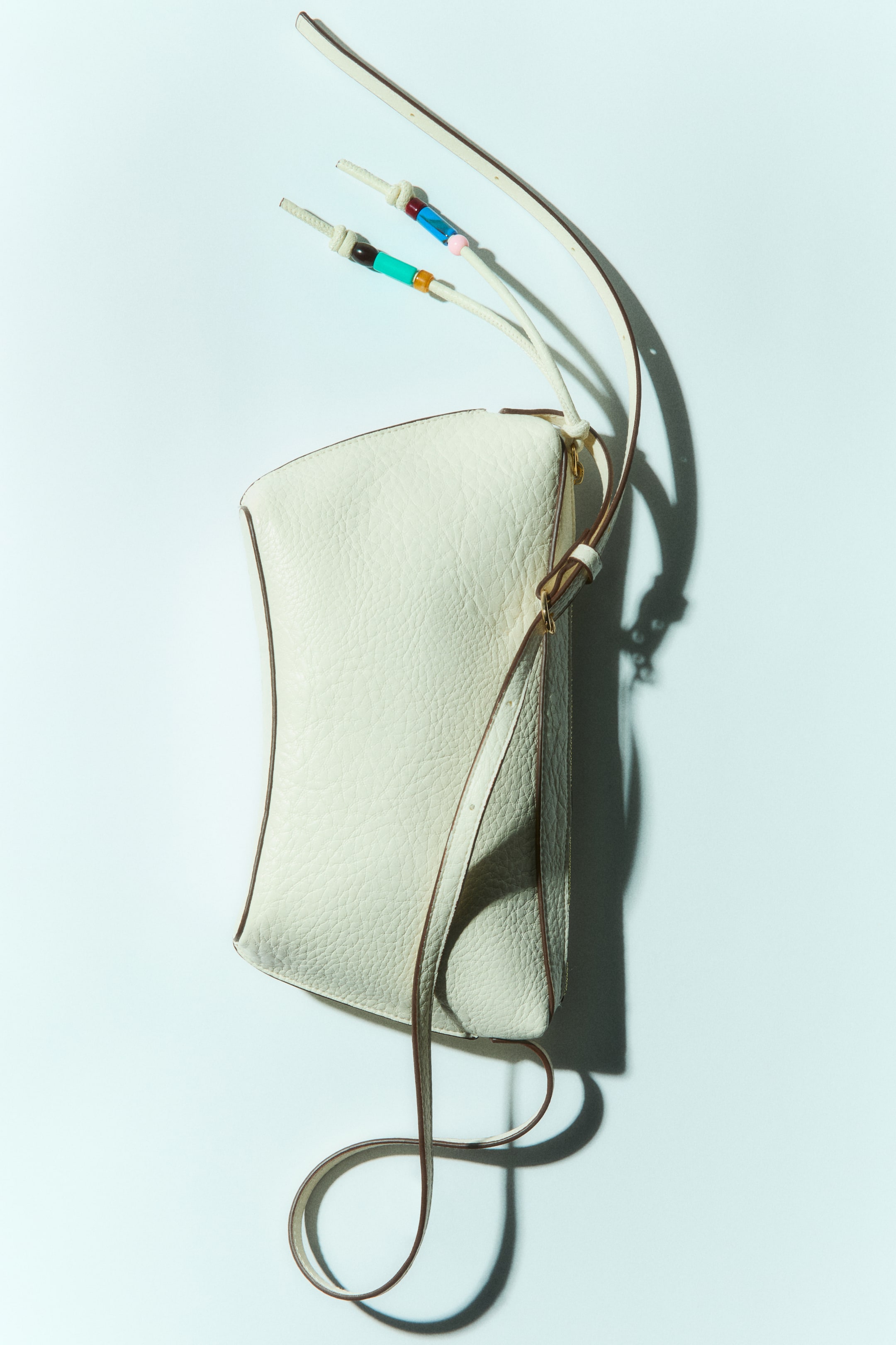 View larger image: An off-white shoulder bag crafted from coated fabric showcases a textured finish and a top zip closure. A lengthy, knotted pull tab adorned with plastic beads in assorted designs extends from the zipper, and an adjustable shoulder strap with a round metal buckle is affixed.