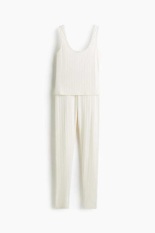 Ein nach vorne gerichtetes, cremefarbenes, geripptes zweiteiliges Loungewear-Set hängt vertikal. Das Set umfasst ein Tanktop mit einem Rundhalsausschnitt mit Spitzenbesatz und einer kleinen Schleife in der Mitte sowie passende, gerippte Hosen in voller Länge. - 1