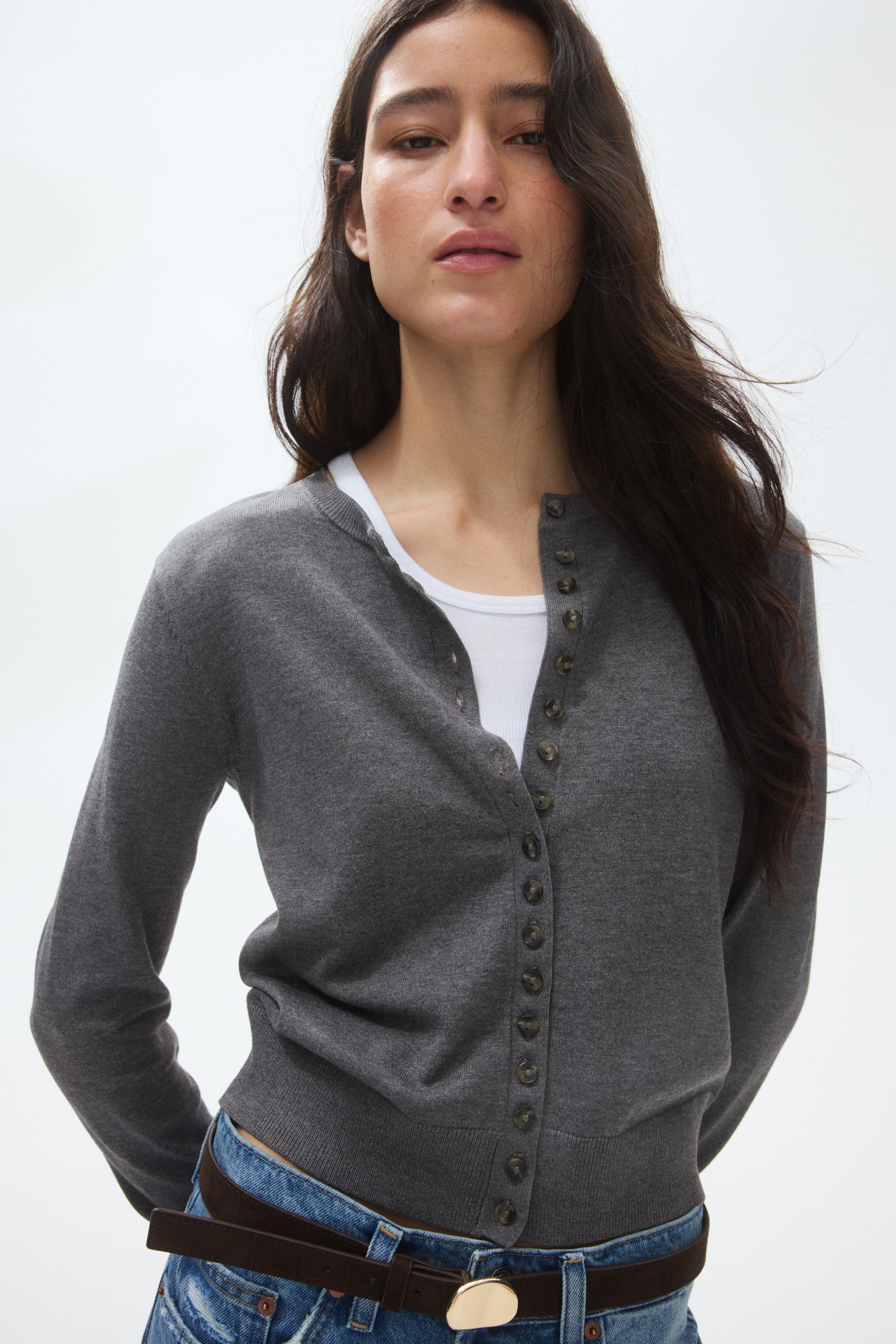 View larger image: A dark grey cardigan features long sleeves, a crew neck, and a full button placket with small dark buttons, slightly angled to the right. It also showcases a ribbed hem and is layered over a white ribbed tank top.