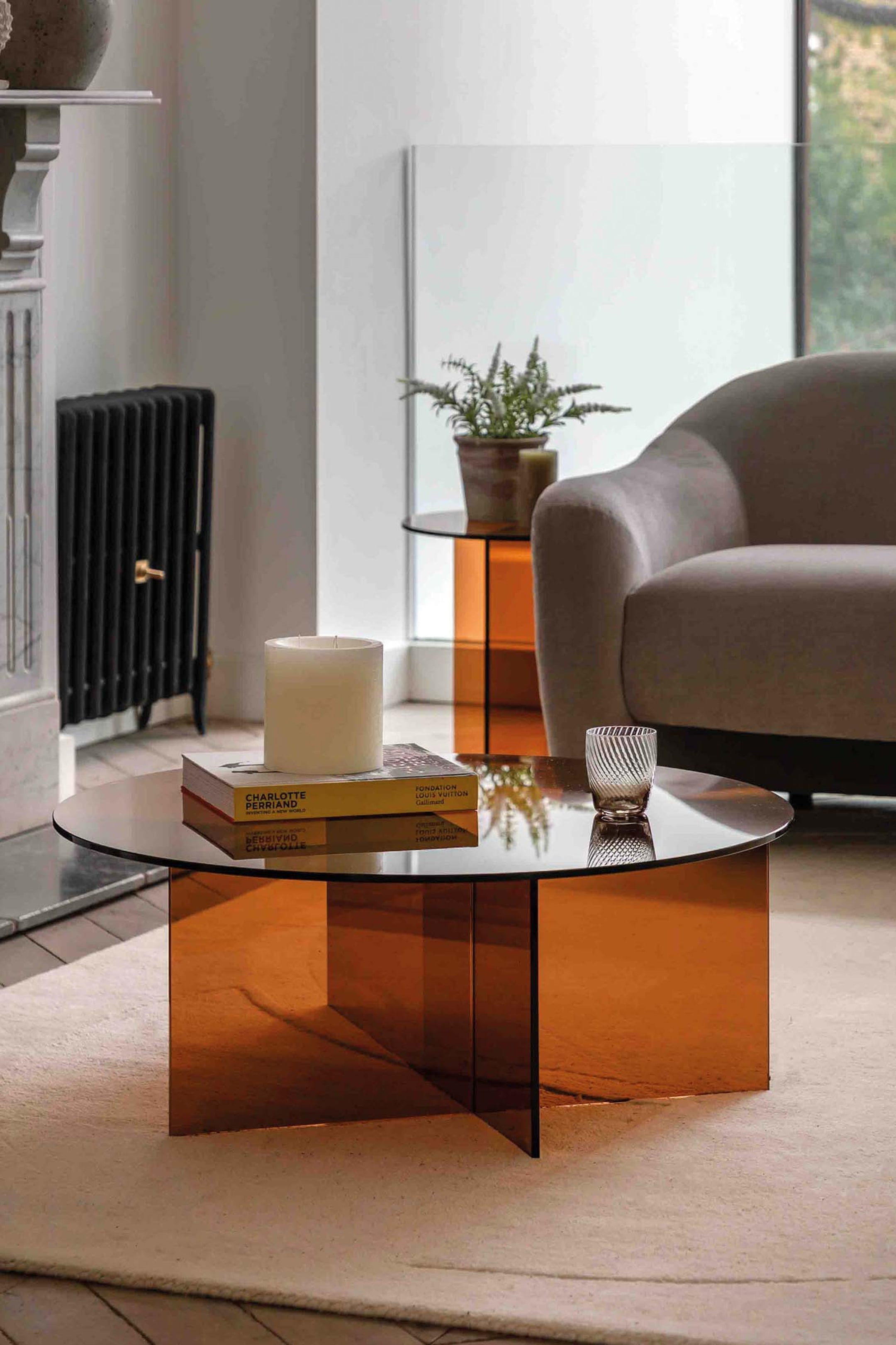 View larger image: An amber-toned round coffee table features a translucent circular top and an X-shaped base, both crafted from the same material. The tabletop displays a stack of books, a cream candle, and a textured glass tumbler.