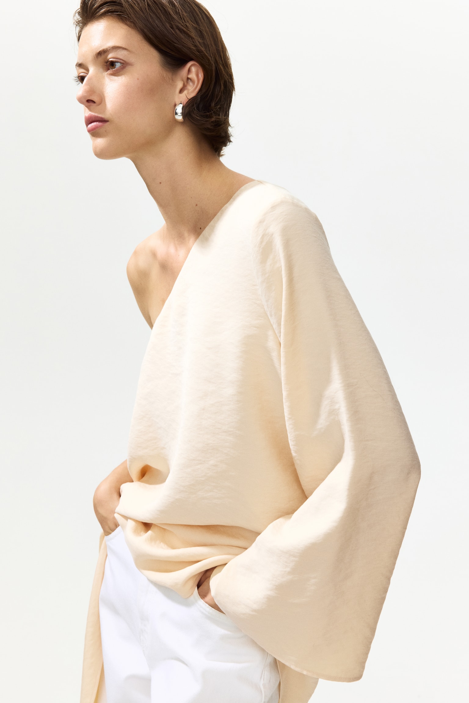 The model is wearing a one-shoulder top crafted from a modal-blend weave. It boasts an asymmetric neckline, a single long sleeve, and an asymmetric hem that drapes gracefully as the model poses with one hand in a trouser pocket. - 1