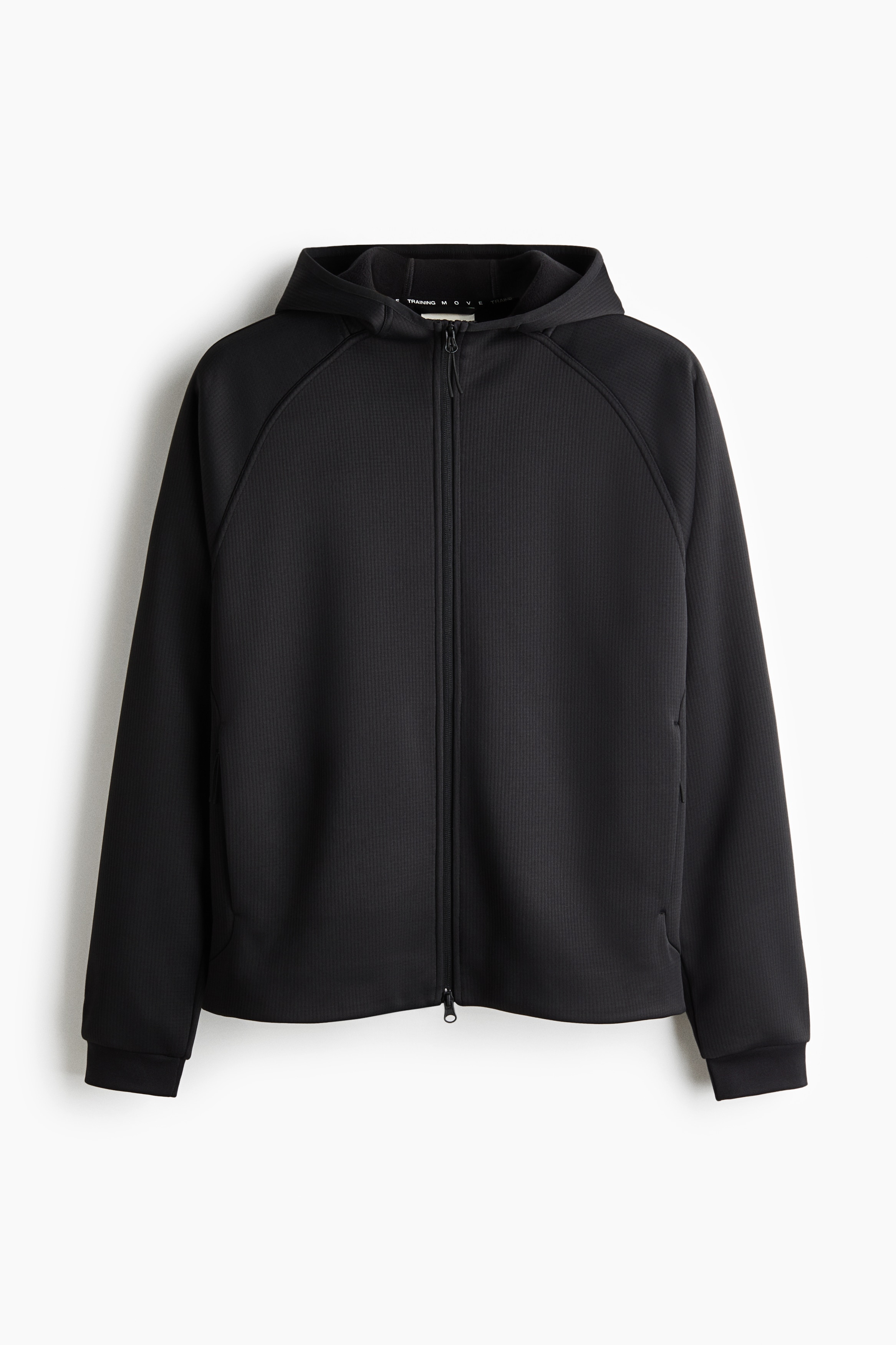 The model is wearing a dark grey, textured zip-up hoodie, fully zipped to the neck over a white collar. The hoodie features raglan sleeves and is styled with black athletic shorts over tights, grey knit gloves and a black beanie.