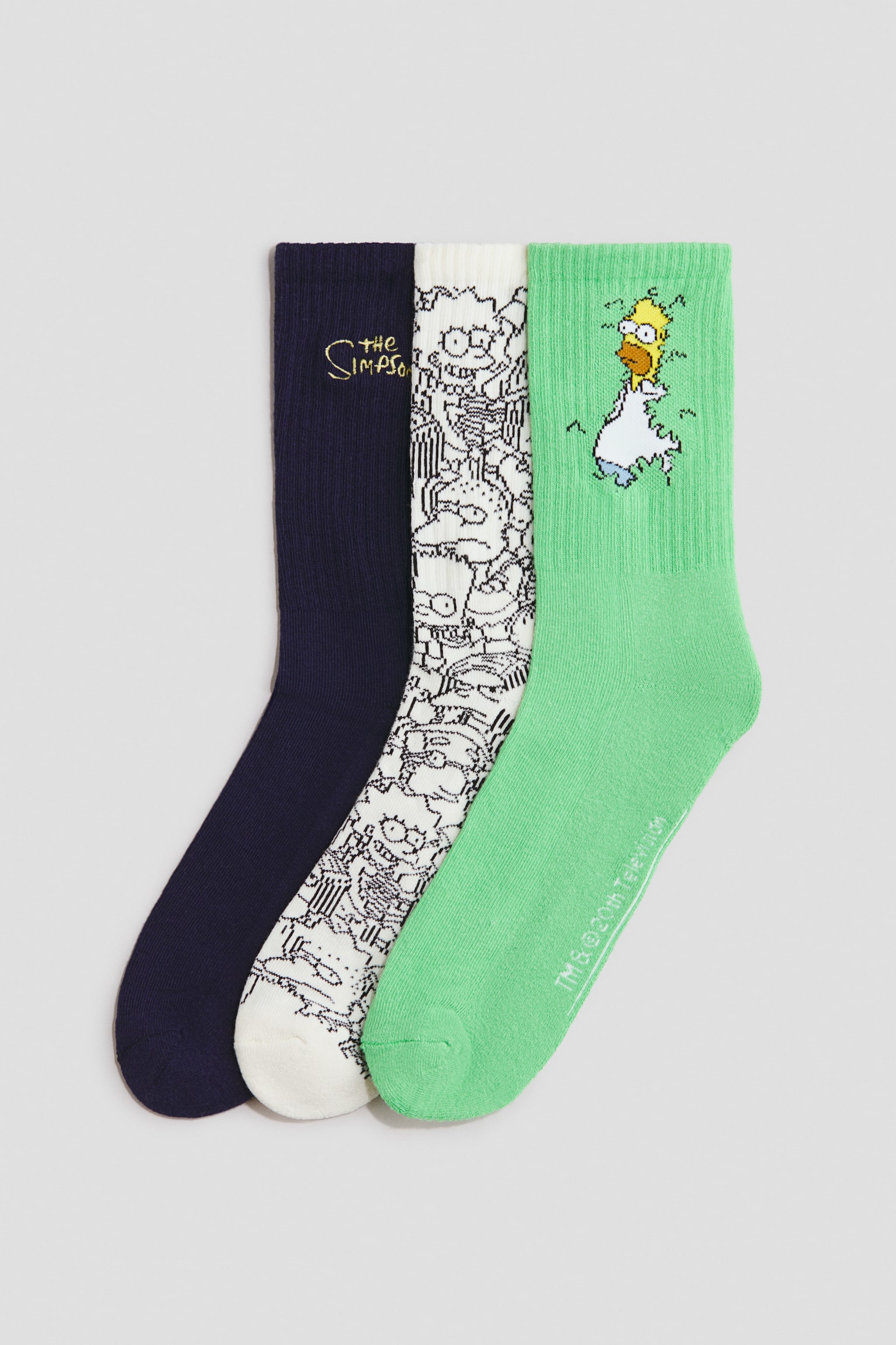 Three crew socks are displayed in a flat lay presentation, each featuring a different The Simpsons-inspired design. The first sock is navy blue with a yellow embroidered logo, the second is white with an all-over black-and-white character print, and the third is green, featuring a graphic of Homer Simpson backing into a bush near the ribbed cuff.