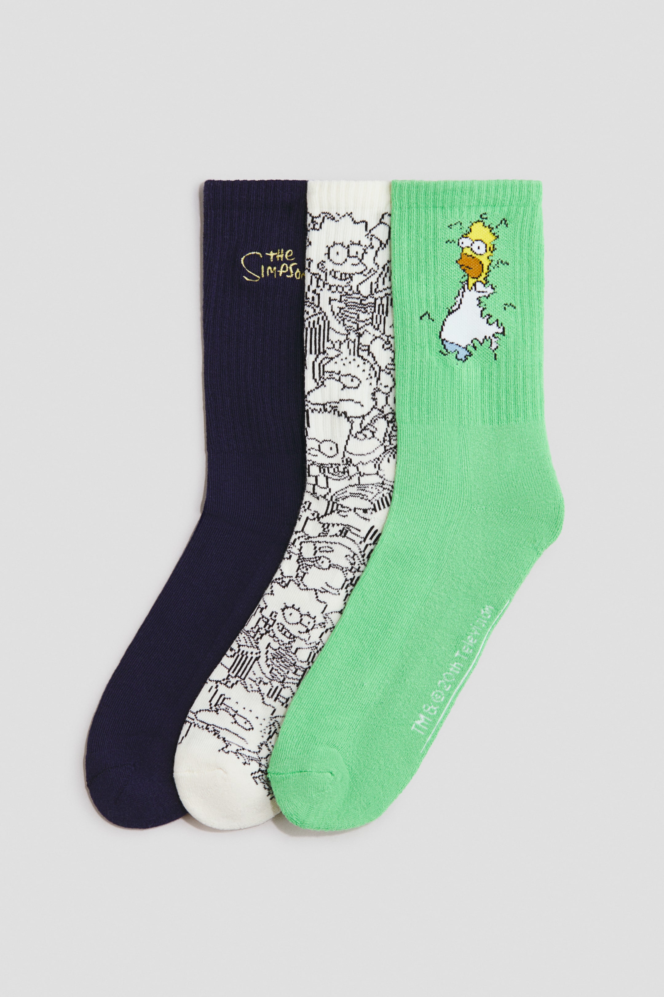 View larger image: Three crew socks are displayed in a flat lay arrangement, each featuring a different The Simpsons-themed design. The first sock is navy blue with a yellow embroidered logo, the second is white with an all-over black-and-white character print, and the third is green, featuring a graphic of Homer Simpson backing into a bush near the ribbed cuff.