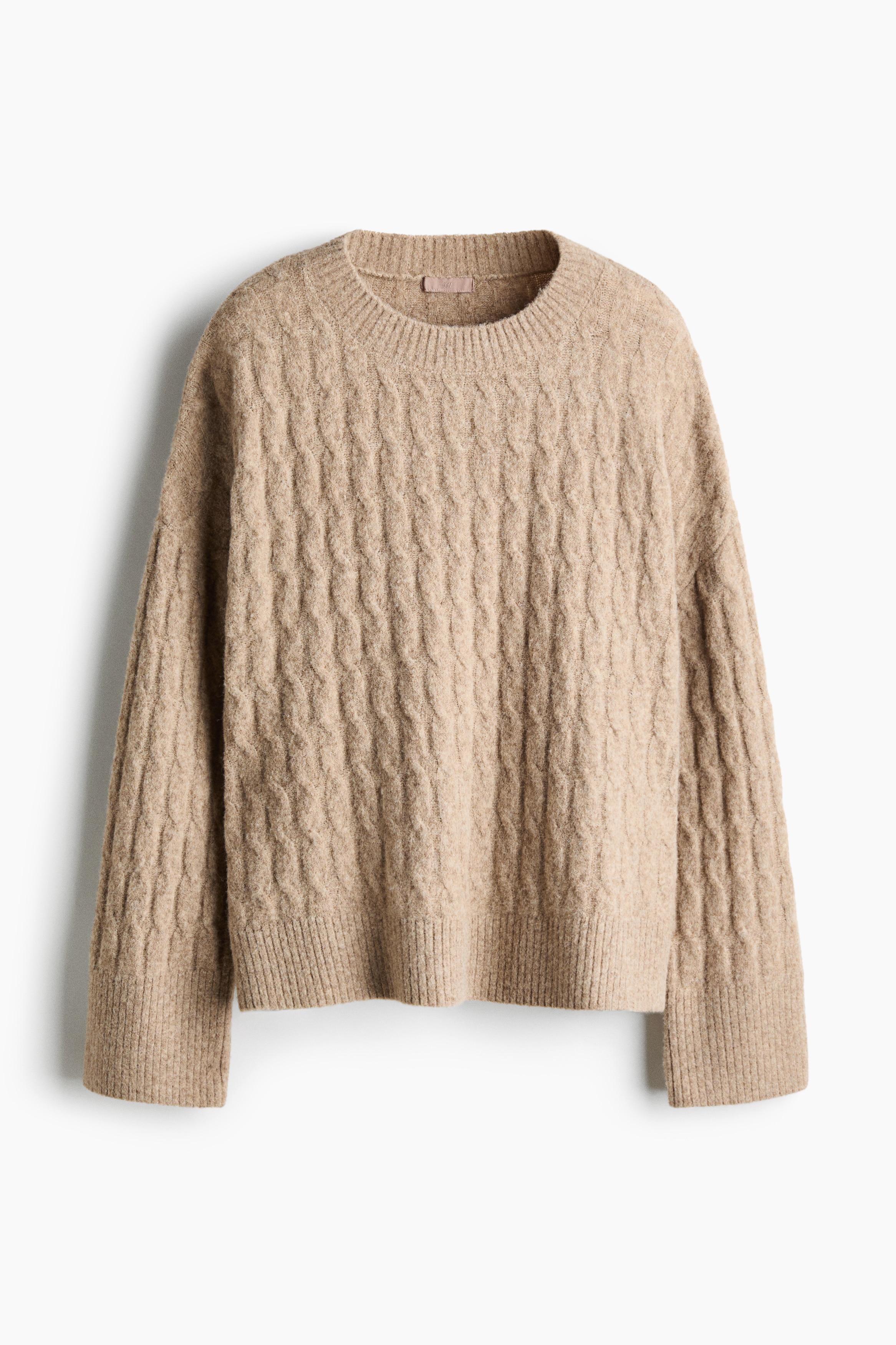 A beige, loose-fit cable-knit jumper is being worn by the model, featuring a ribbed crew neckline and dropped shoulders. The jumper is layered over a white long-sleeve top and paired with matching cable-knit trousers as the model sits.