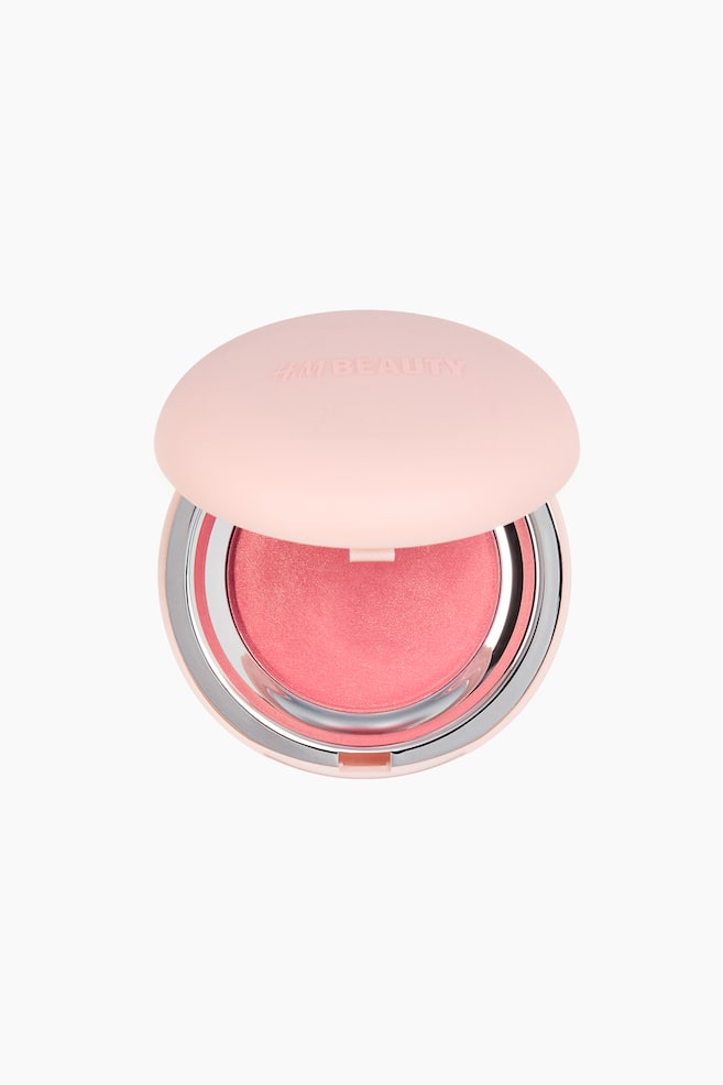 Een ronde blusher-compact, met de voorkant naar voren gericht (FRONT), bevat een koraalroze blusher met een subtiele glans. Het matte lichtroze dekseltje staat iets open, waardoor de blusherpan en een zilverkleurige binnenrand zichtbaar zijn, terwijl "H&M BEAUTY" in reliëf op het deksel staat. - 1