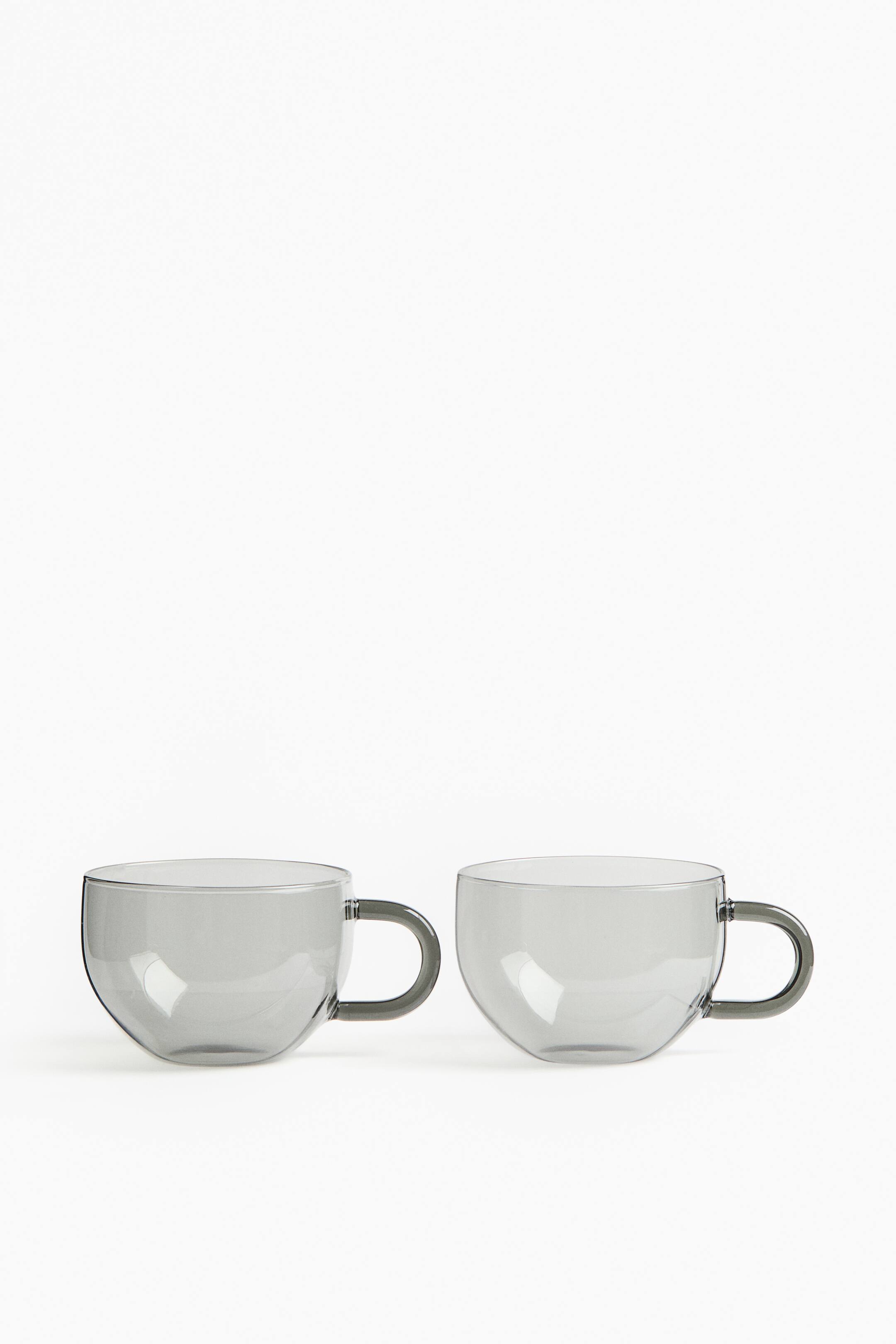 View larger image: Two transparent, grey-tinted glass cups are standing side-by-side, each featuring a wide, rounded body and a thick D-shaped handle.