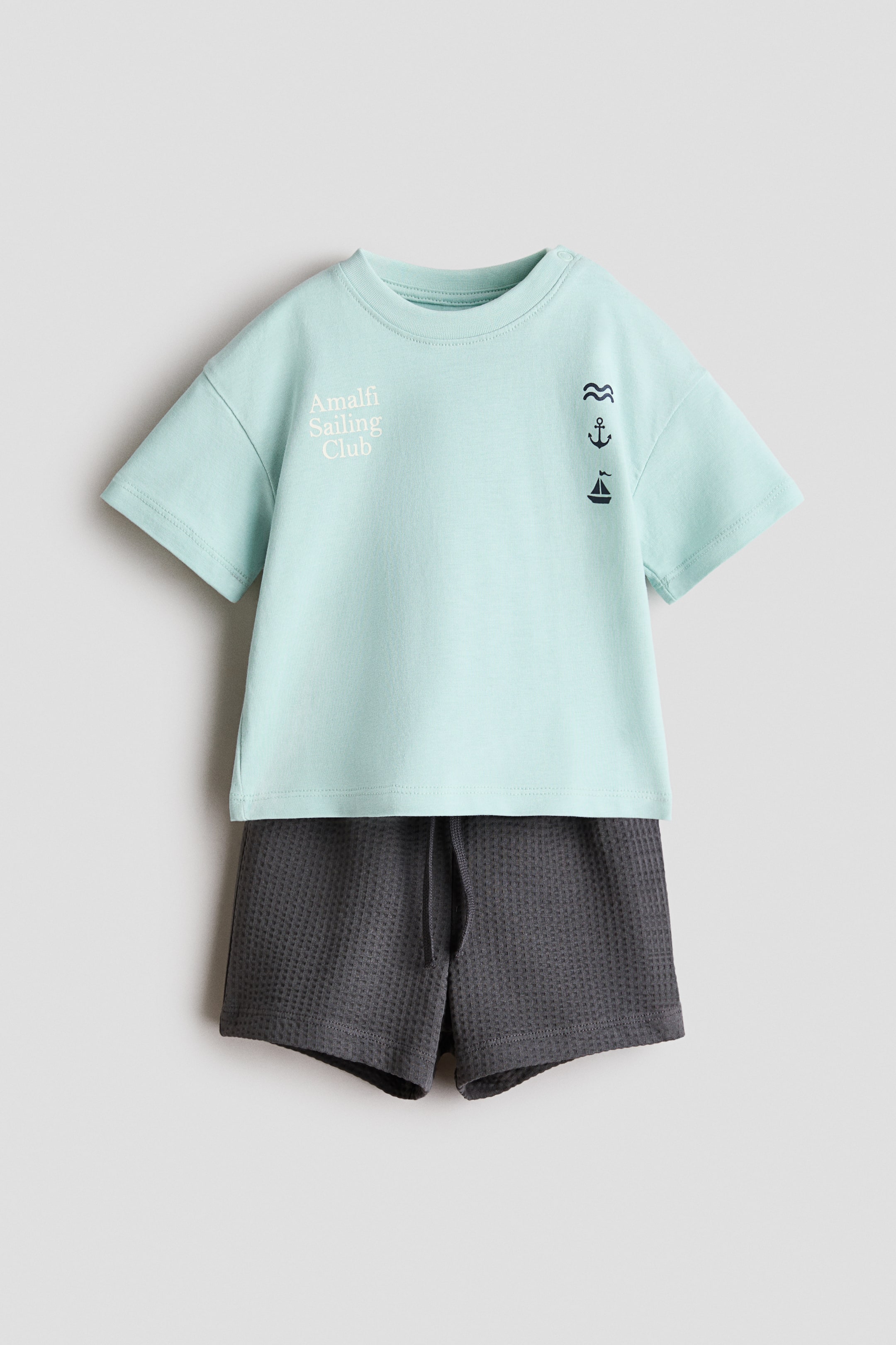 View larger image: A 2-piece cotton jersey set is presenting its front, featuring a light blue short-sleeved T-shirt with a ribbed crew neck and dark grey waffle knit shorts. The T-shirt is displaying "Amalfi Sailing Club" text on the left chest and stacked wave, anchor, and sailboat icons on the right, while the shorts are showing a drawstring waist.