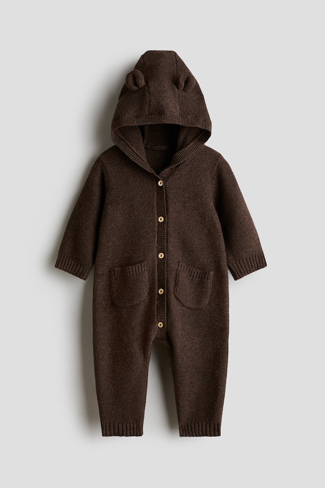 Dieser dunkelbraune Strickoverall aus Baumwolle zeigt eine frontale Ansicht mit einer Kapuze mit kleinen gestrickten Ohren. Er verfÃ¼gt Ã¼ber einen zentralen Knopfverschluss mit hellen KnÃ¶pfen, zwei aufgesetzte Taschen vorne sowie RippbÃ¼ndchen an den Ãrmeln und KnÃ¶cheln. - 1