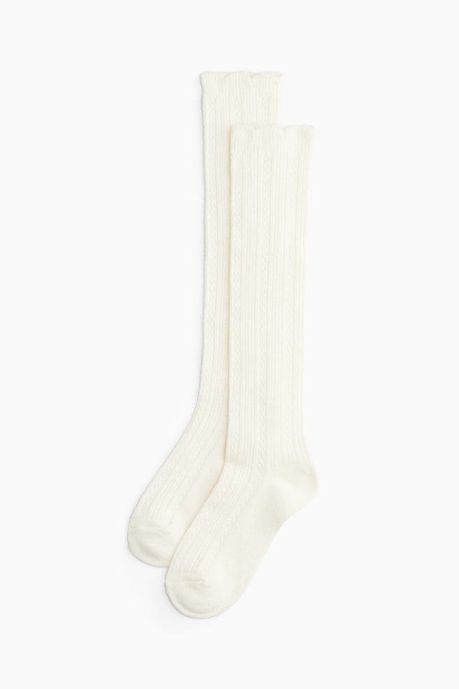 Cremefarbene knielange Socken aus weichem, gemustertem Strick liegen flach in frontaler Ansicht und zeigen ein vertikales Rippen- und Zopfstrickmuster. Die Socken verfÃ¼gen oben Ã¼ber elastische, gewellte BÃ¼ndchen mit ausgeformter Ferse und Zehenpartie. - 1