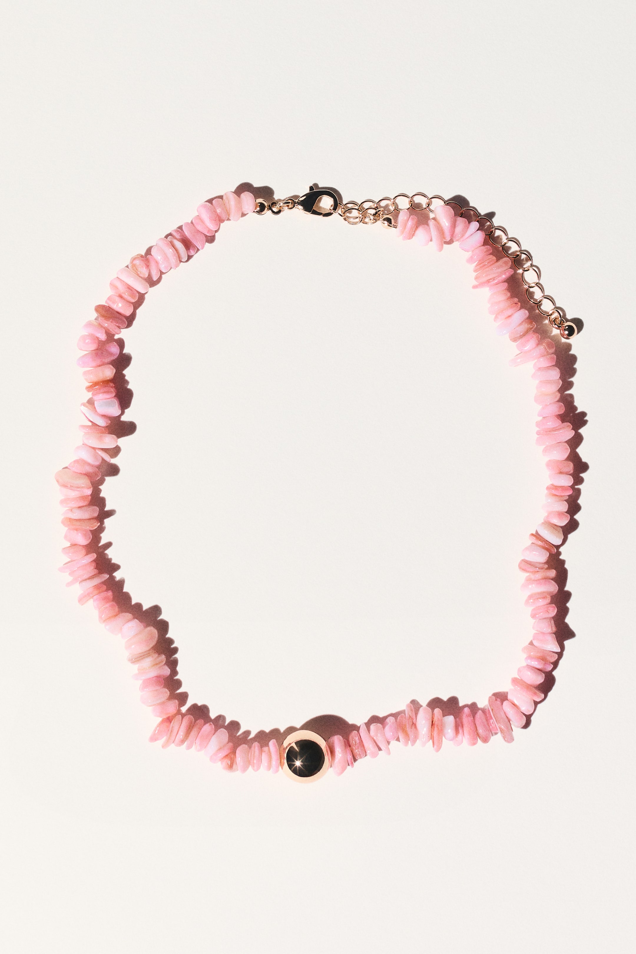 Größeres Bild ansehen: Eine Halskette aus unregelmäßig geformten rosafarbenen Splitterperlen liegt flach und bildet eine offene Kurve. Sie verfügt über einen runden, goldfarbenen Anhänger mit schwarzem Zentrum und einem subtilen Funkeln, und ein goldfarbener Karabinerverschluss mit Verlängerungskette ist sichtbar.