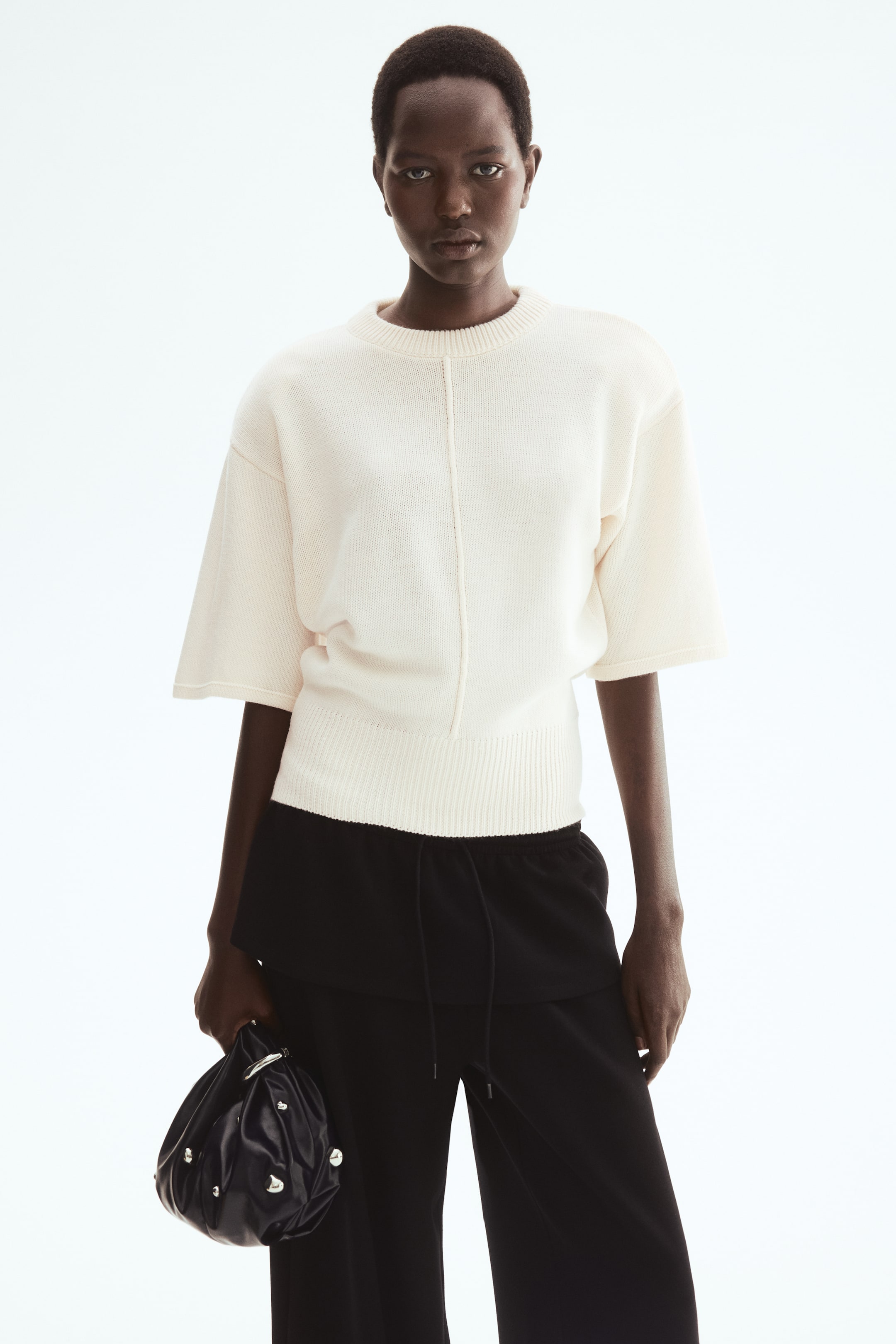 查看大圖: 模特穿著一件奶油色針織上衣,正面有連接縫線,落肩設計和短袖。領口、袖口和下擺均可見羅紋設計。