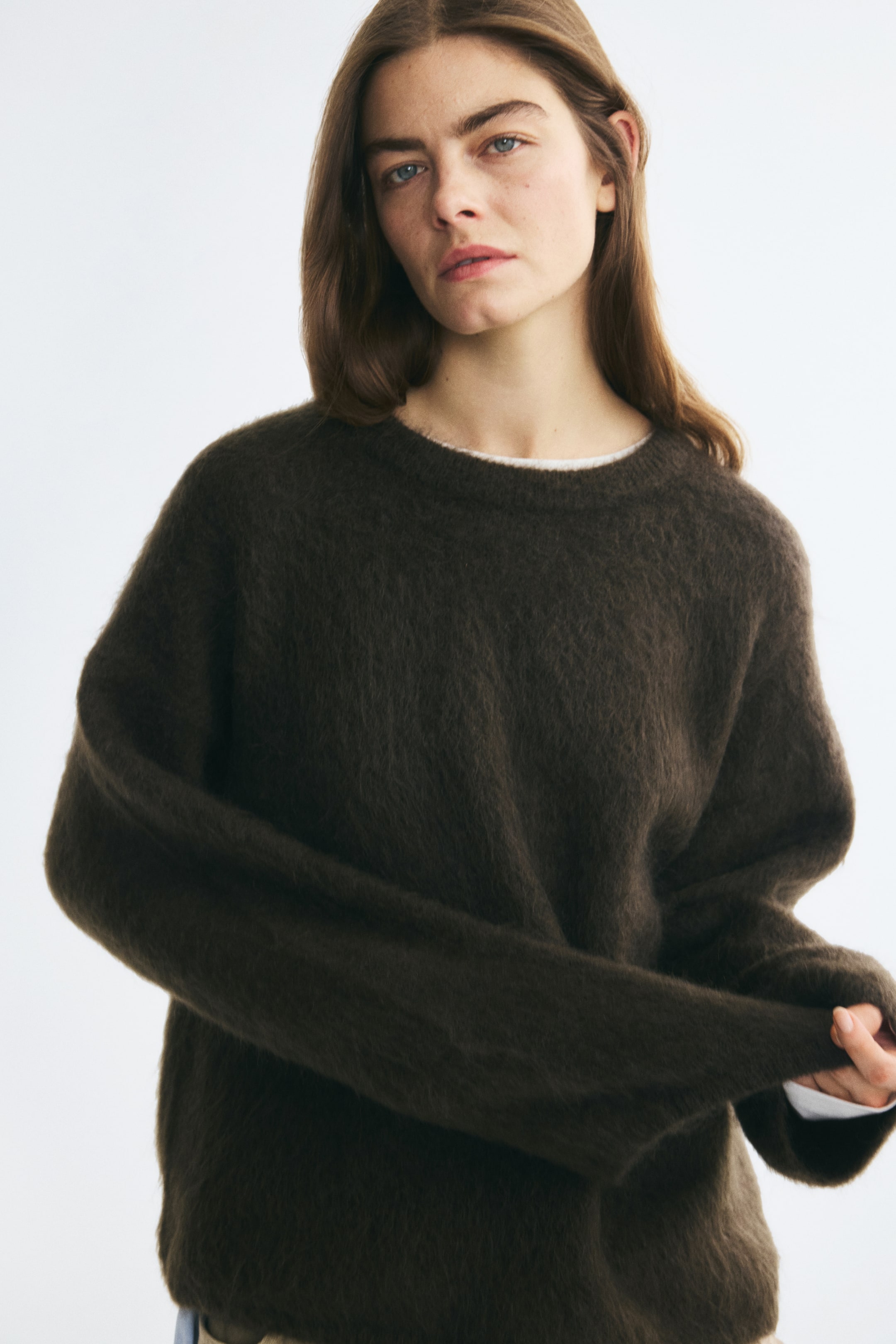 View larger image: An oversized, dark brown fuzzy knit sweater is being worn by the model, who is holding the fabric of one long sleeve. The sweater features dropped shoulders, a ribbed crew neckline, and is layered over a white top visible at the collar and cuff.