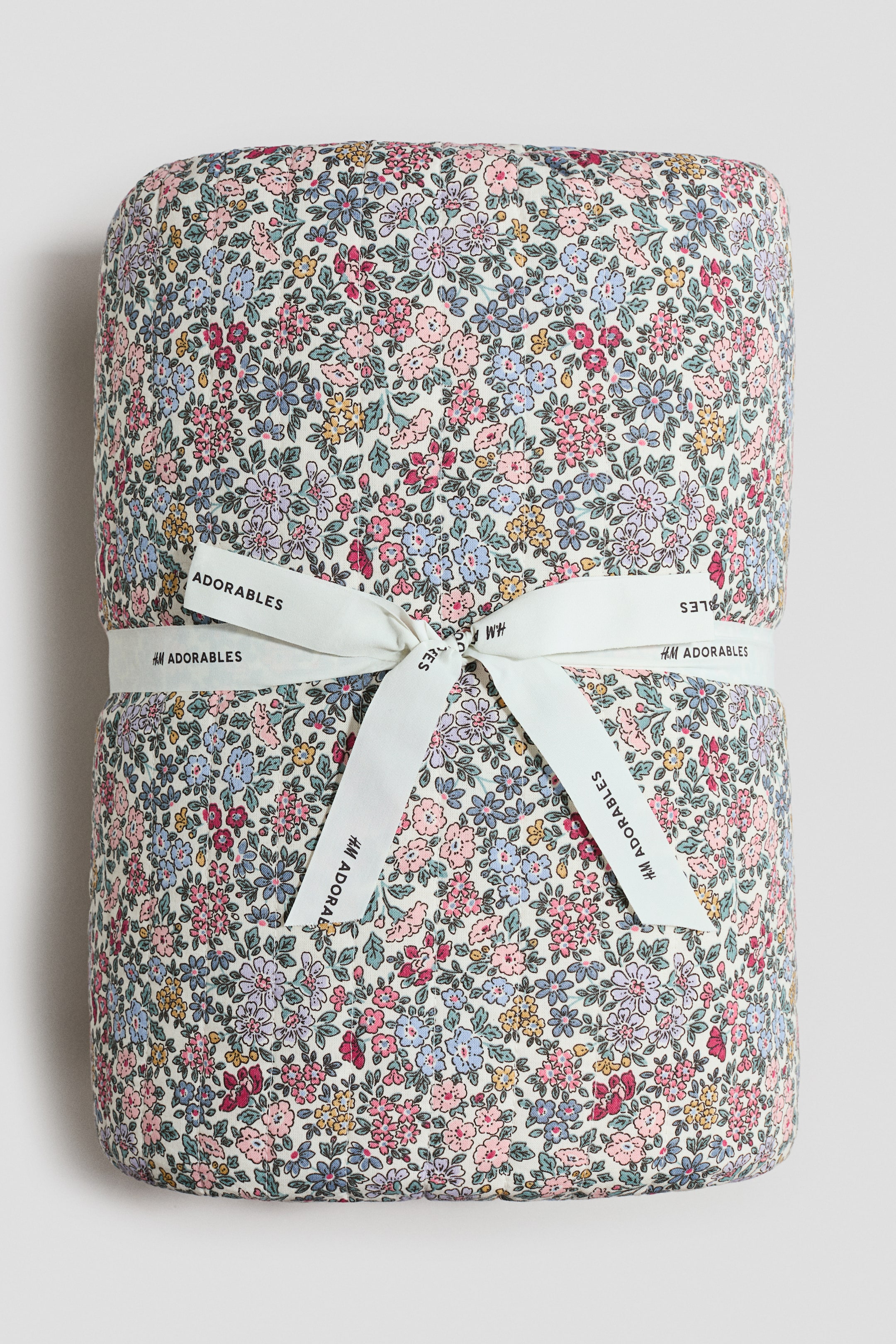 Agrandir l'image: Une couverture matelassée affiche un imprimé floral vif à petite échelle en rose, bleu, violet et jaune. Un ruban blanc, imprimé avec "H&M ADORABLES," est enroulé autour de la couverture et noué en un nœud à l'avant.