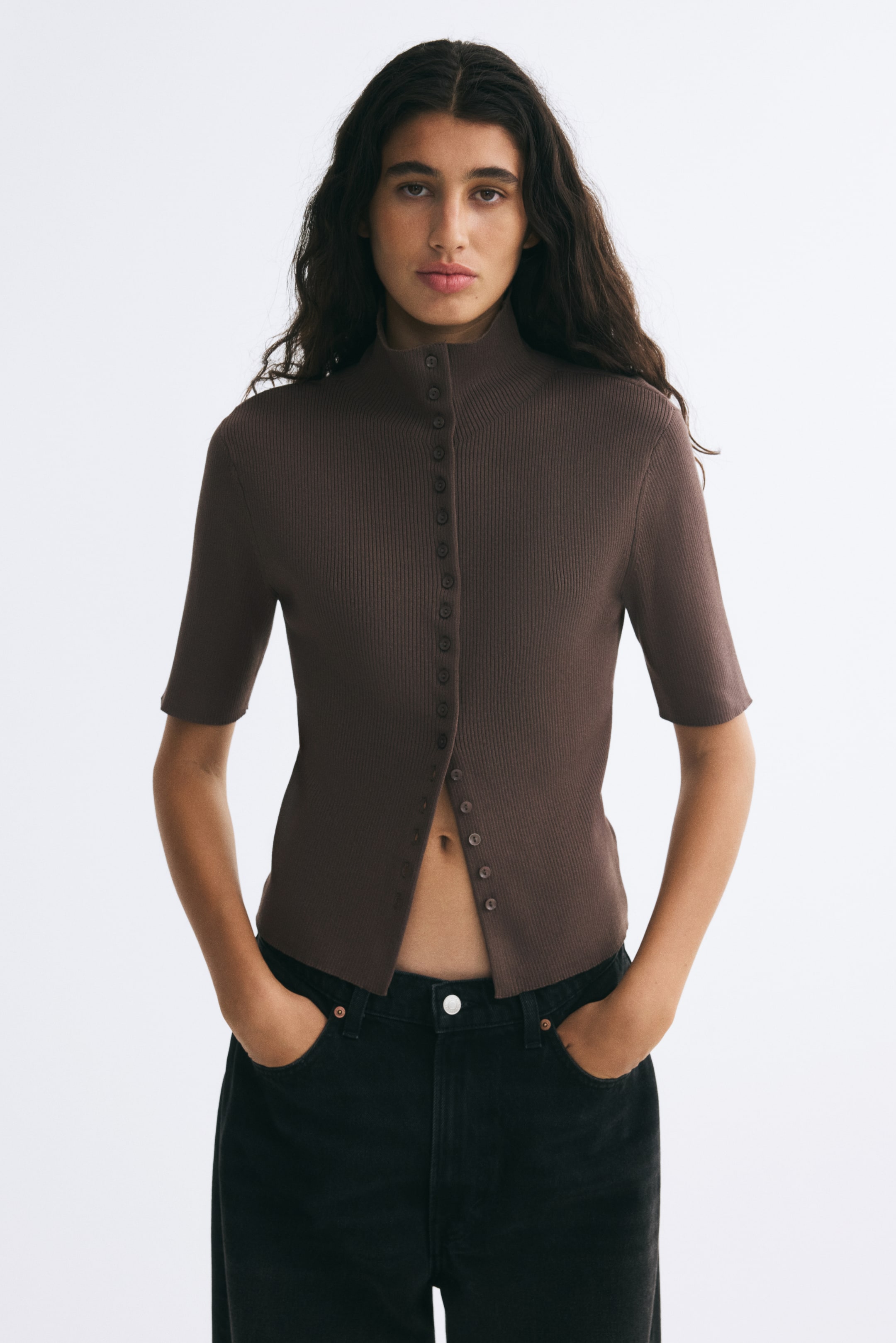 View larger image: The model is wearing a brown, fitted rib-knit cardigan with a high neck, short sleeves, and a button-front closure. The cardigan is left partially unbuttoned at the hem and styled with black jeans.