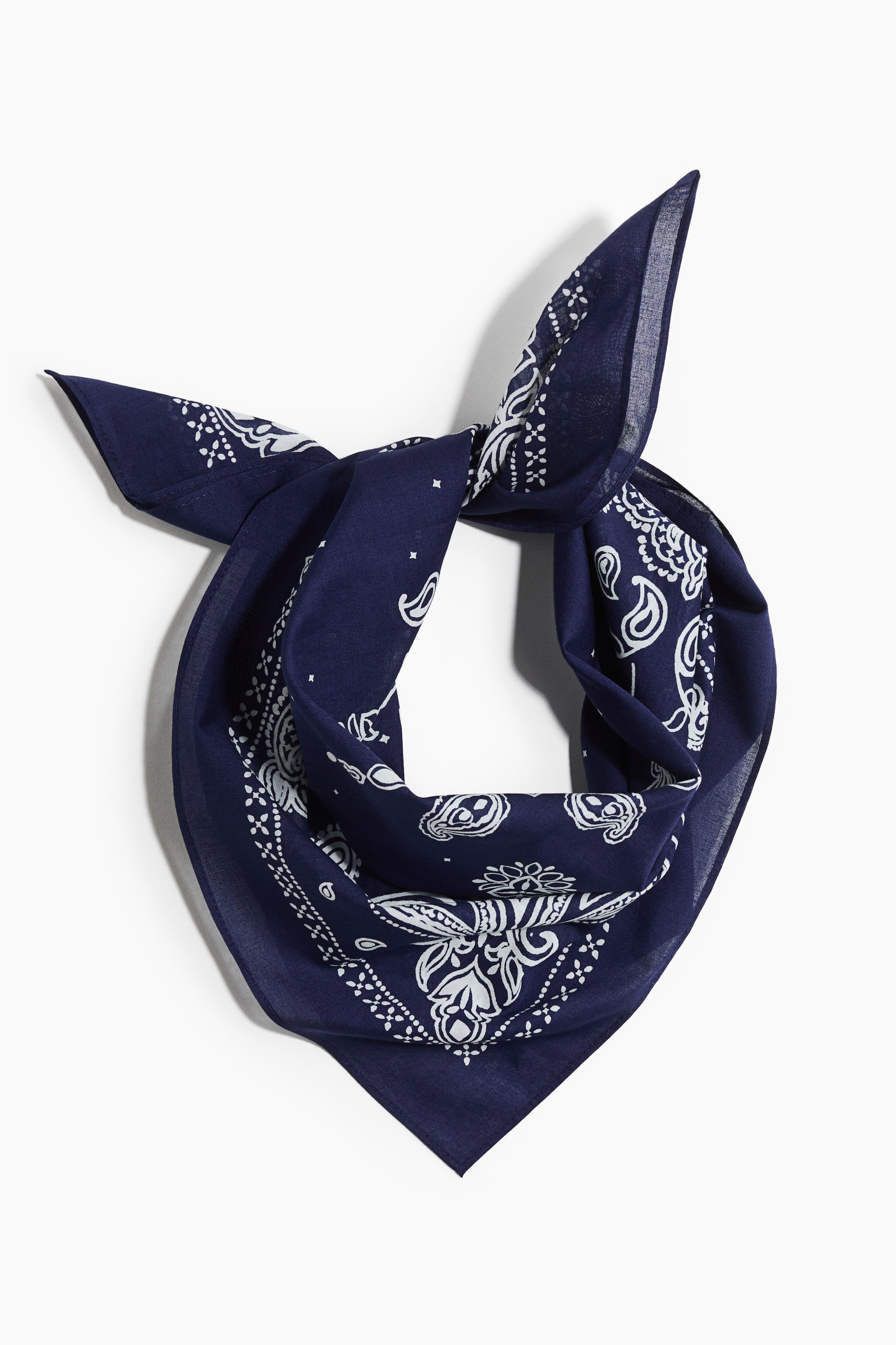 Visa större bild: Halsduk i ett mönstrat bomullstyg visar en marinblå färg med vita paisley- och geometriska motiv. Den är knuten upptill, vilket bildar en knut, och presenterar en triangulär form med två spetsiga ändar som pekar framåt.