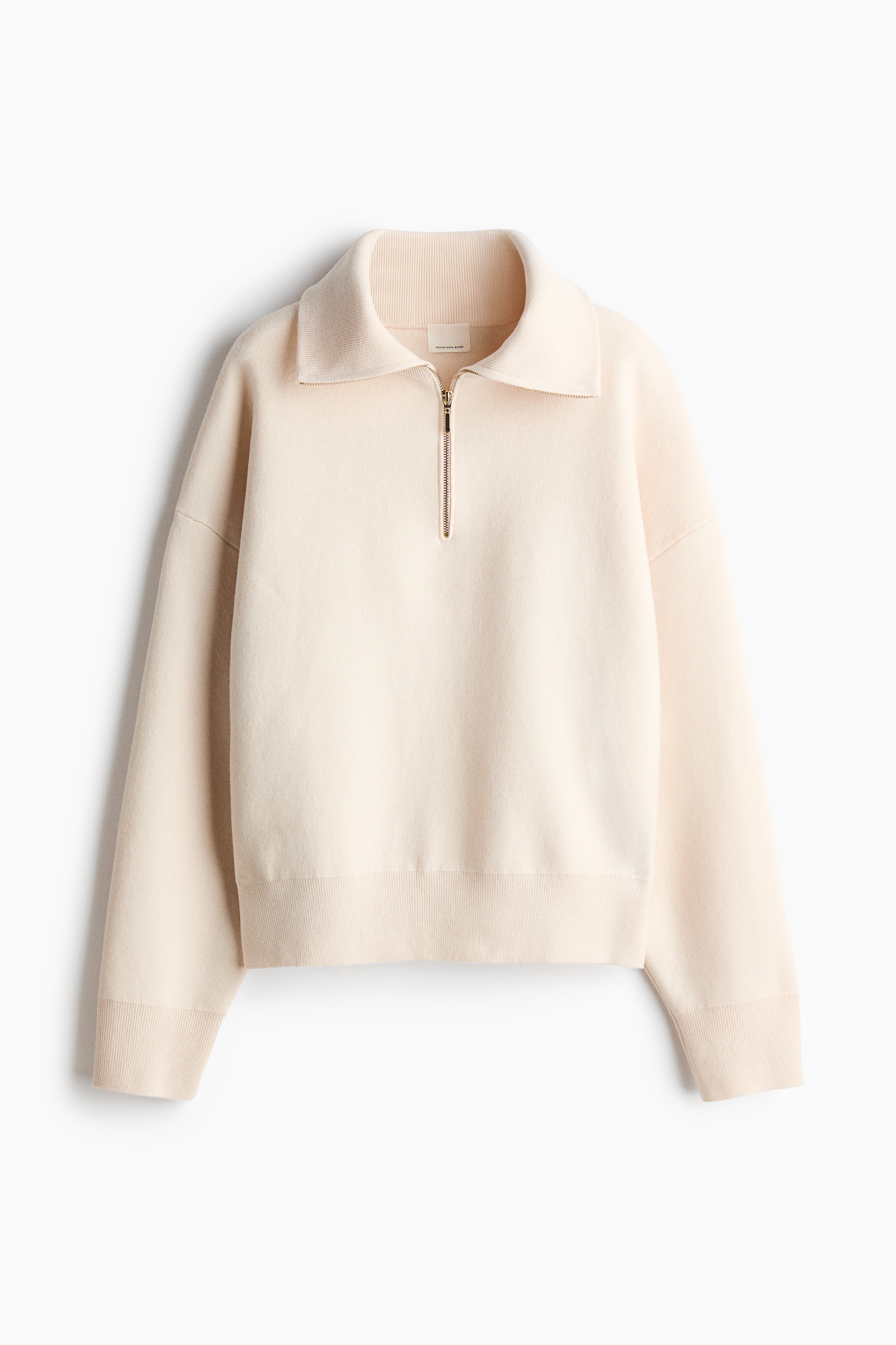 An off-white, fine-knit sweater featuring a high ribbed funnel collar and a zipped-up half-zip with a gold pull tab is being worn by the model. The side-profile view highlights the sweater's dropped shoulder and relaxed, oversized fit.
