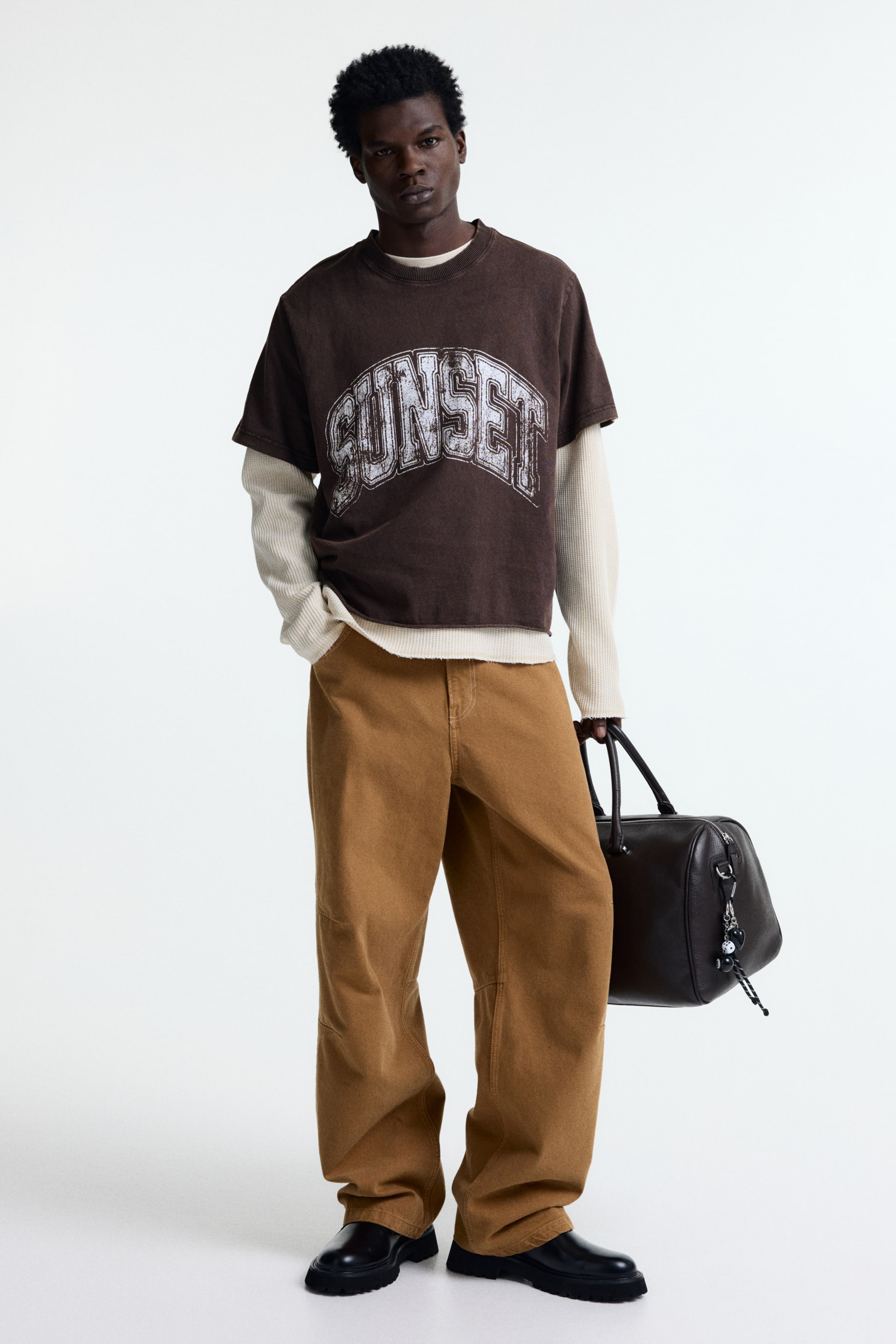View larger image: The model is wearing a brown cotton jersey T-shirt with a regular fit, showcasing a distressed 'SUNSET' print and a round, ribbed neckline. This piece is layered over a light-coloured, long-sleeved top.