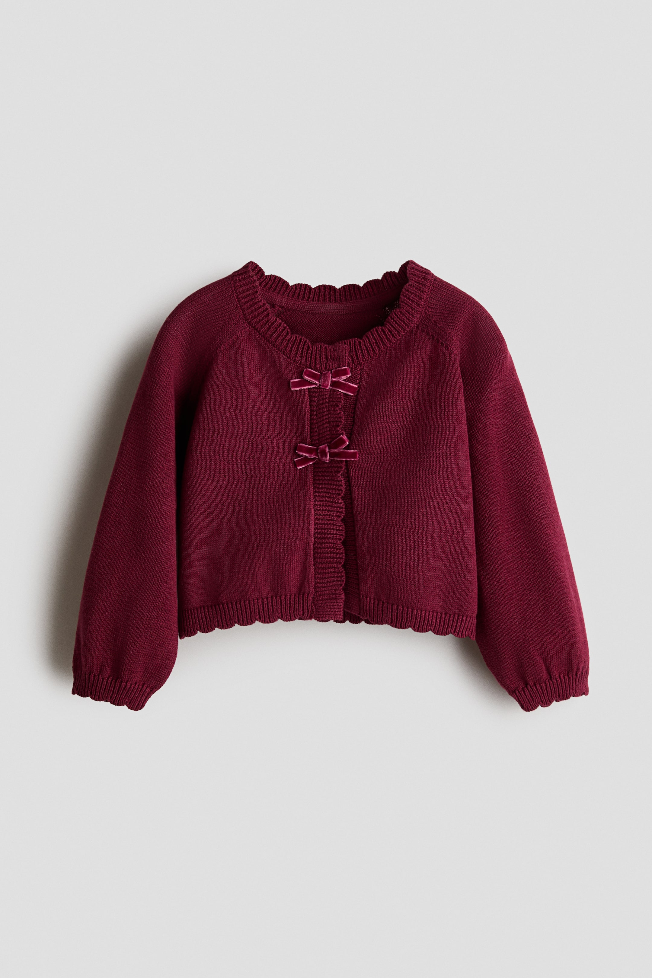 View larger image: A burgundy fine-knit cotton cardigan is shown front-facing, featuring a round neckline, long sleeves, and scalloped ribbed edges along the collar, cuffs, hem, and placket. The garment has a concealed button-down front accented with two small velvet bows near the top.