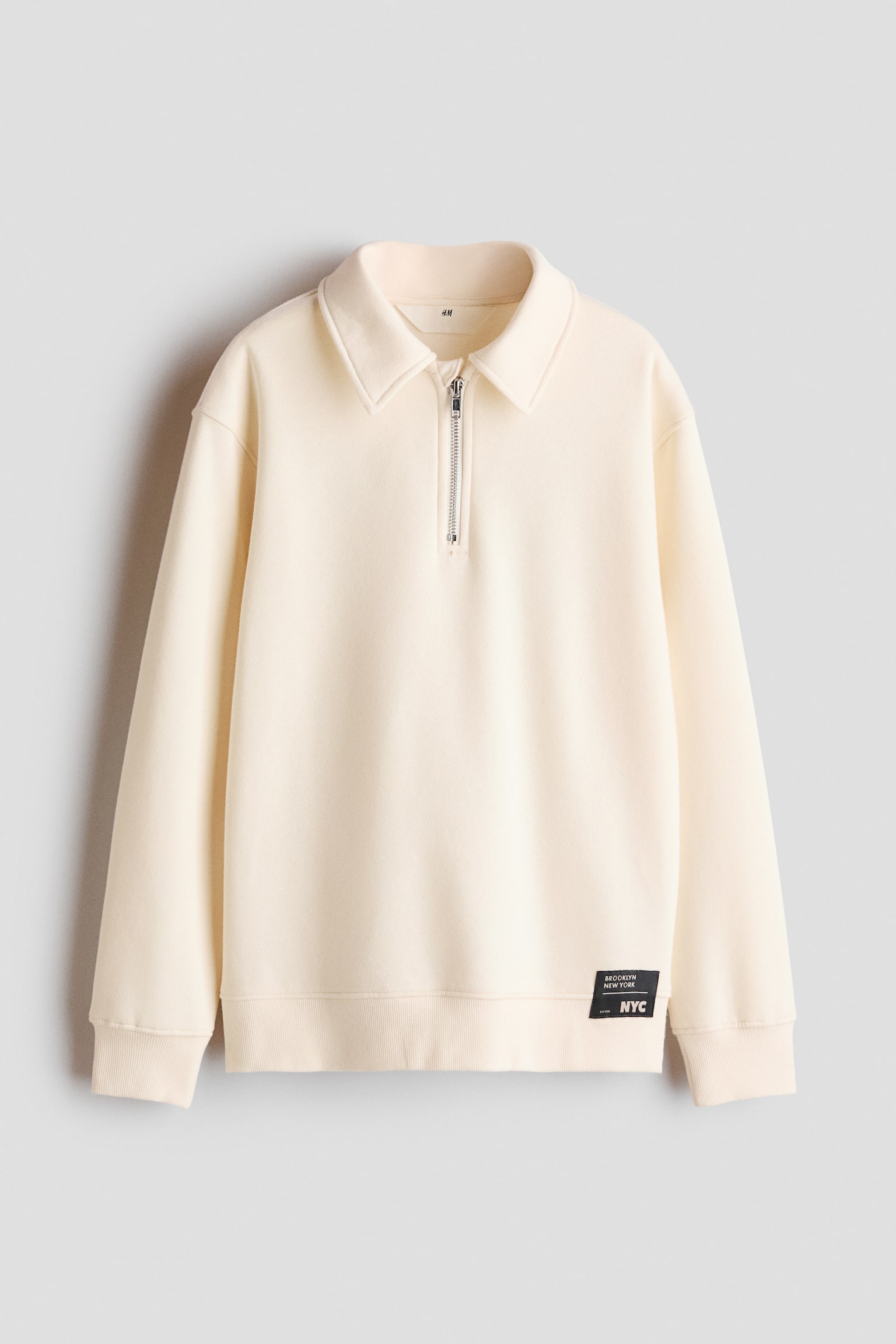 Un sweat-shirt couleur crÃ¨me est prÃ©sentÃ© de face, mettant en valeur un col Ã  pointes avec une fermeture demi-zippÃ©e argentÃ©e. Le vÃªtement prÃ©sente des Ã©paules tombantes, des poignets cÃ´telÃ©s, un bas cÃ´telÃ© et une petite Ã©cusson rectangulaire noir prÃ¨s de lâourlet infÃ©rieur gauche.