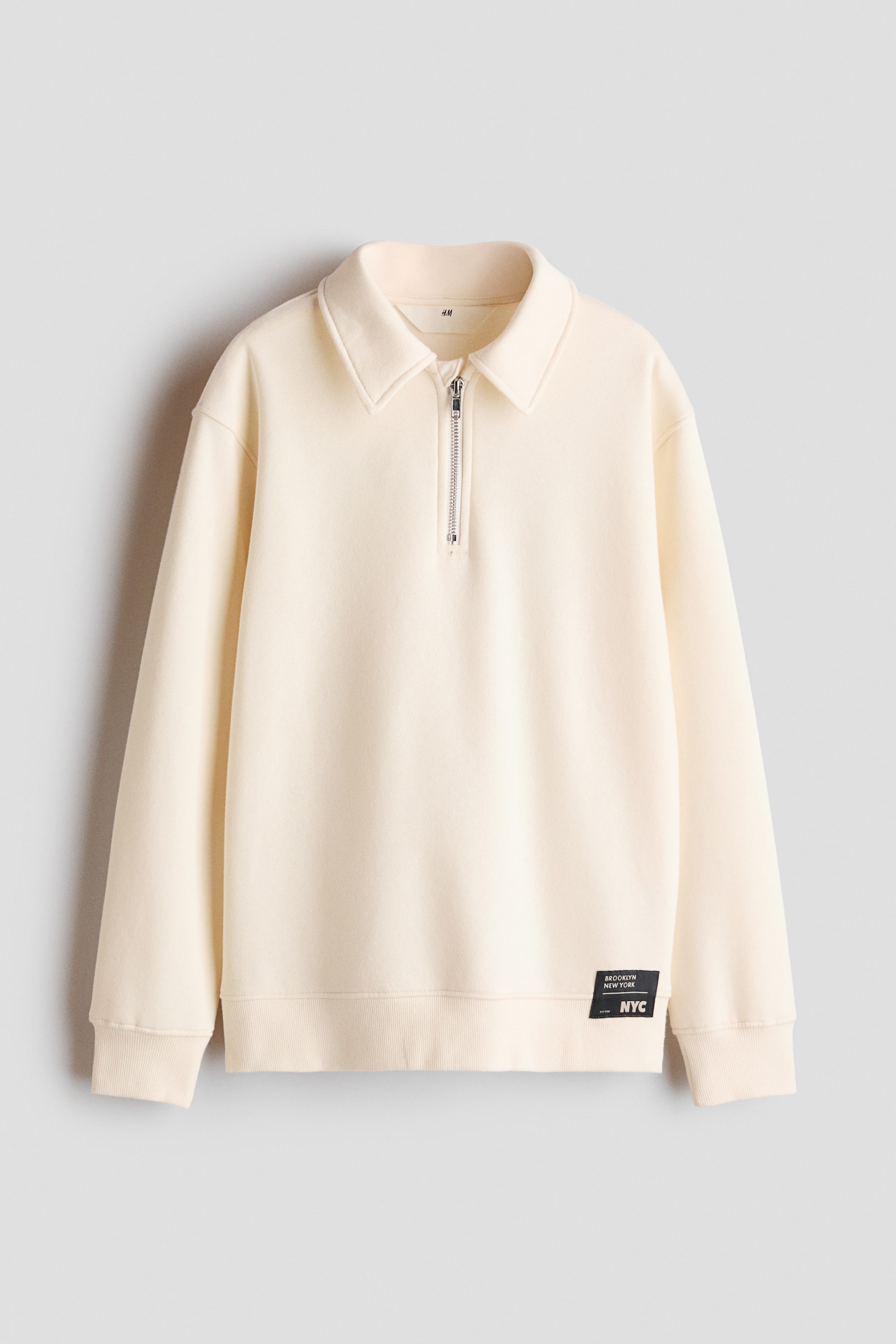 View larger image: A cream-colored sweatshirt is shown from the front, featuring a spread collar with a silver half-zip closure. The garment has dropped shoulders, ribbed cuffs, a ribbed hem, and a small black rectangular patch near the lower left hem.