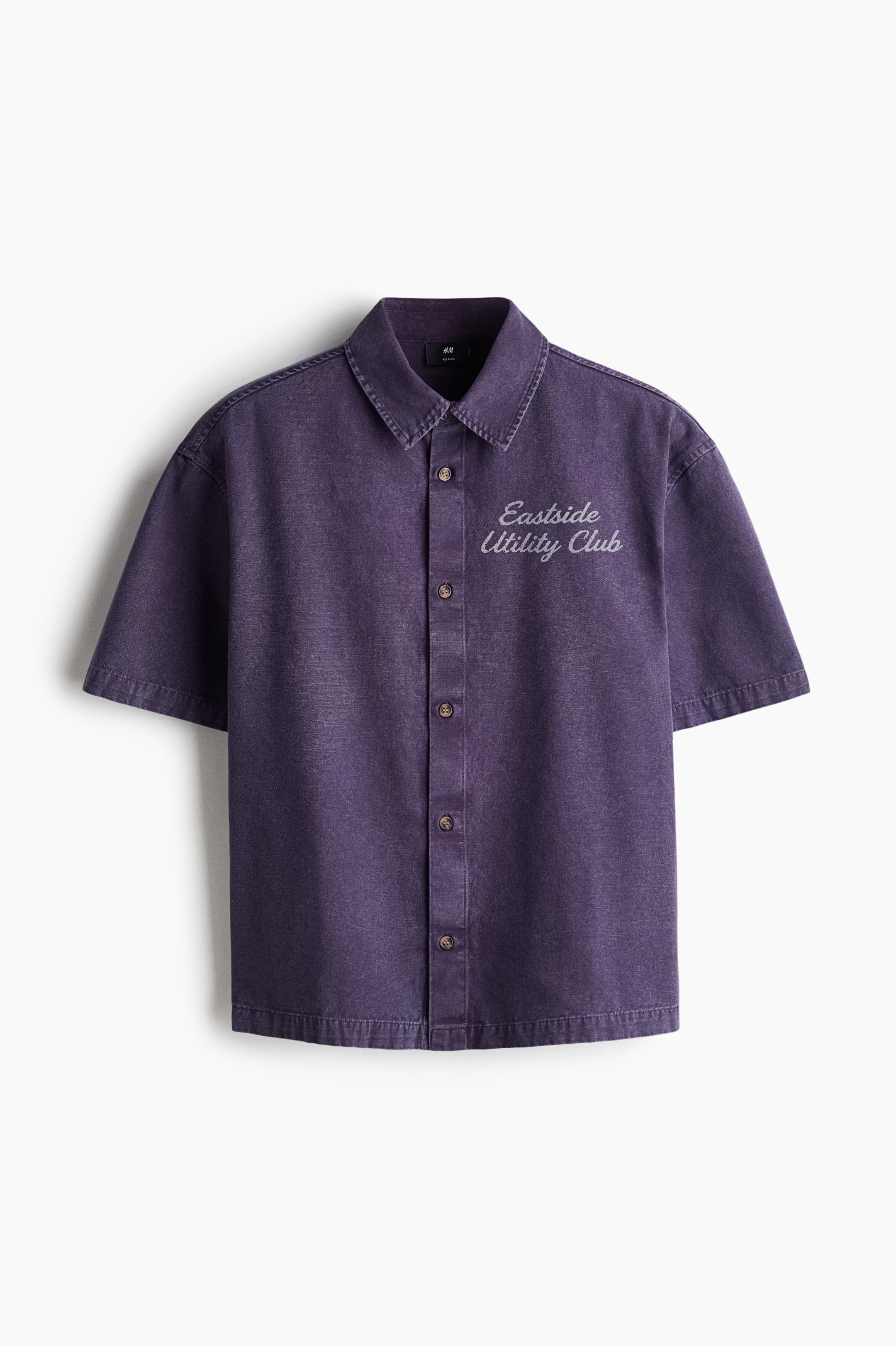 Voir une image plus grande: Une chemise à manches courtes mauve délavé est présentée de face, avec un col à pointes ouvertes et une fermeture boutonnée complète sur le devant avec des boutons foncés. Un texte brodé blanc indiquant « Eastside Utility Club » apparaît sur la poitrine gauche.