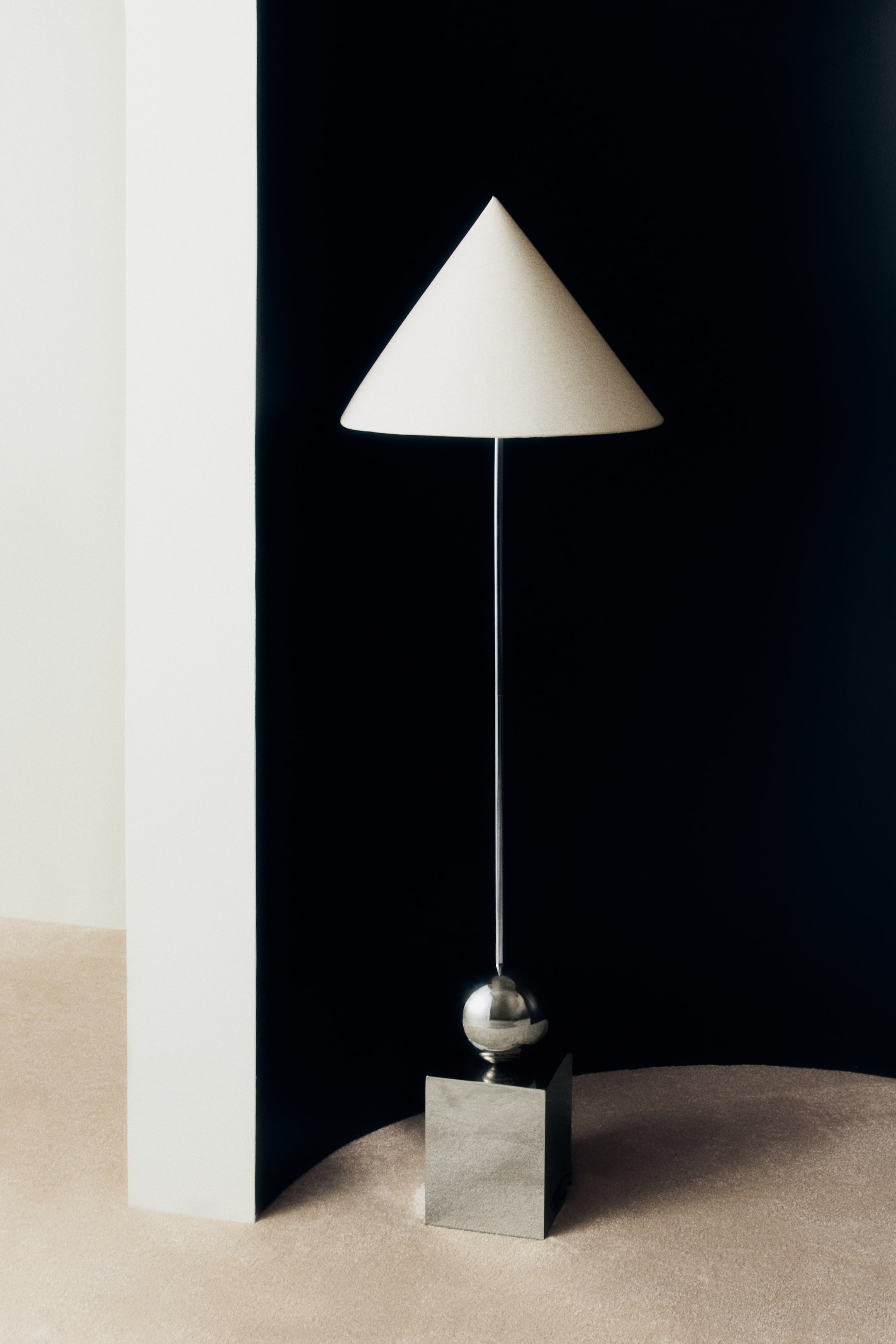 Visualizza immagine più grande: Una lampada da terra vista frontalmente è in posizione eretta, con un design geometrico caratterizzato da un paralume a cono color bianco sporco e da un sottile stelo in metallo lucido. La base in metallo lucido è composta da una sfera metallica appoggiata su un cubo solido.