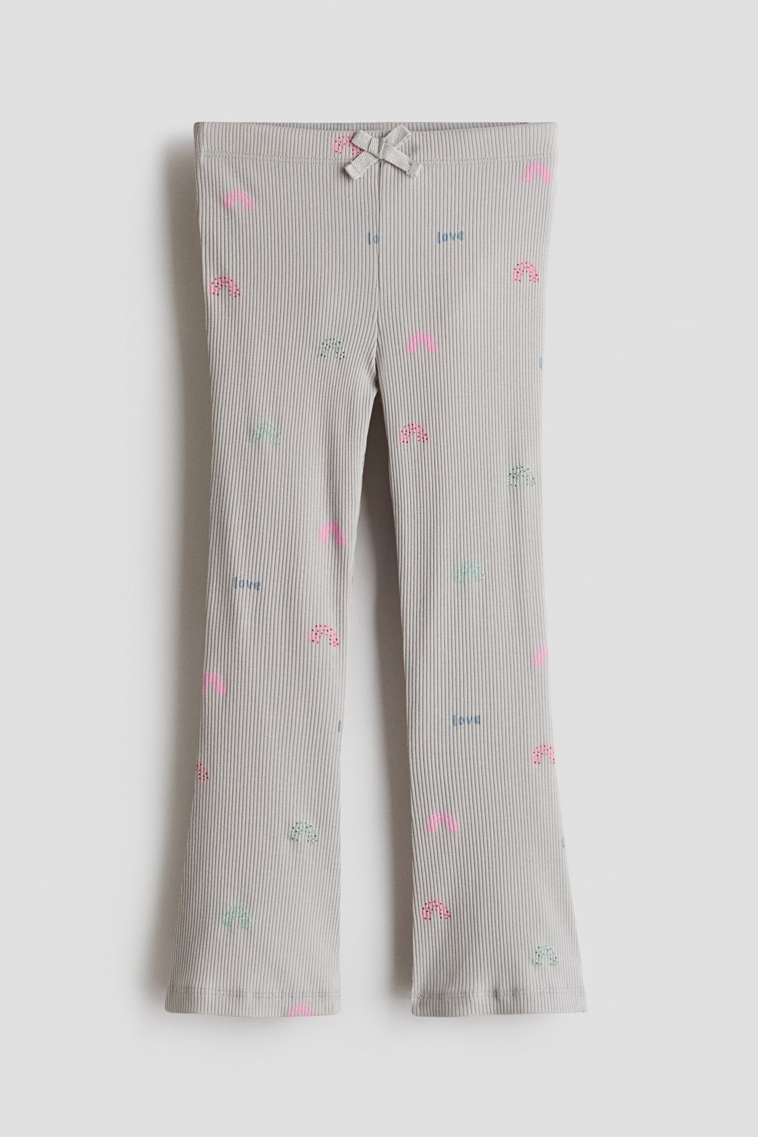 These ribbed cotton jersey leggings are shown from the front, featuring a light grey fabric adorned with an all-over pattern of small pastel rainbows and the word "love". The design includes an elasticated waistband detailed with a small fabric bow at the centre front, and slightly flared legs ending in wavy lettuce hems.