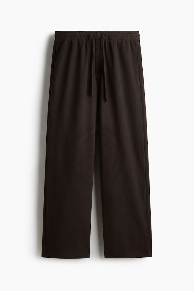 Un pantalon brun foncé présente une vue de face, avec une texture côtelée verticale fine. Le pantalon est conçu avec une taille élastiquée à cordon de serrage, des poches latérales dans les coutures et une coupe droite et décontractée. - 1