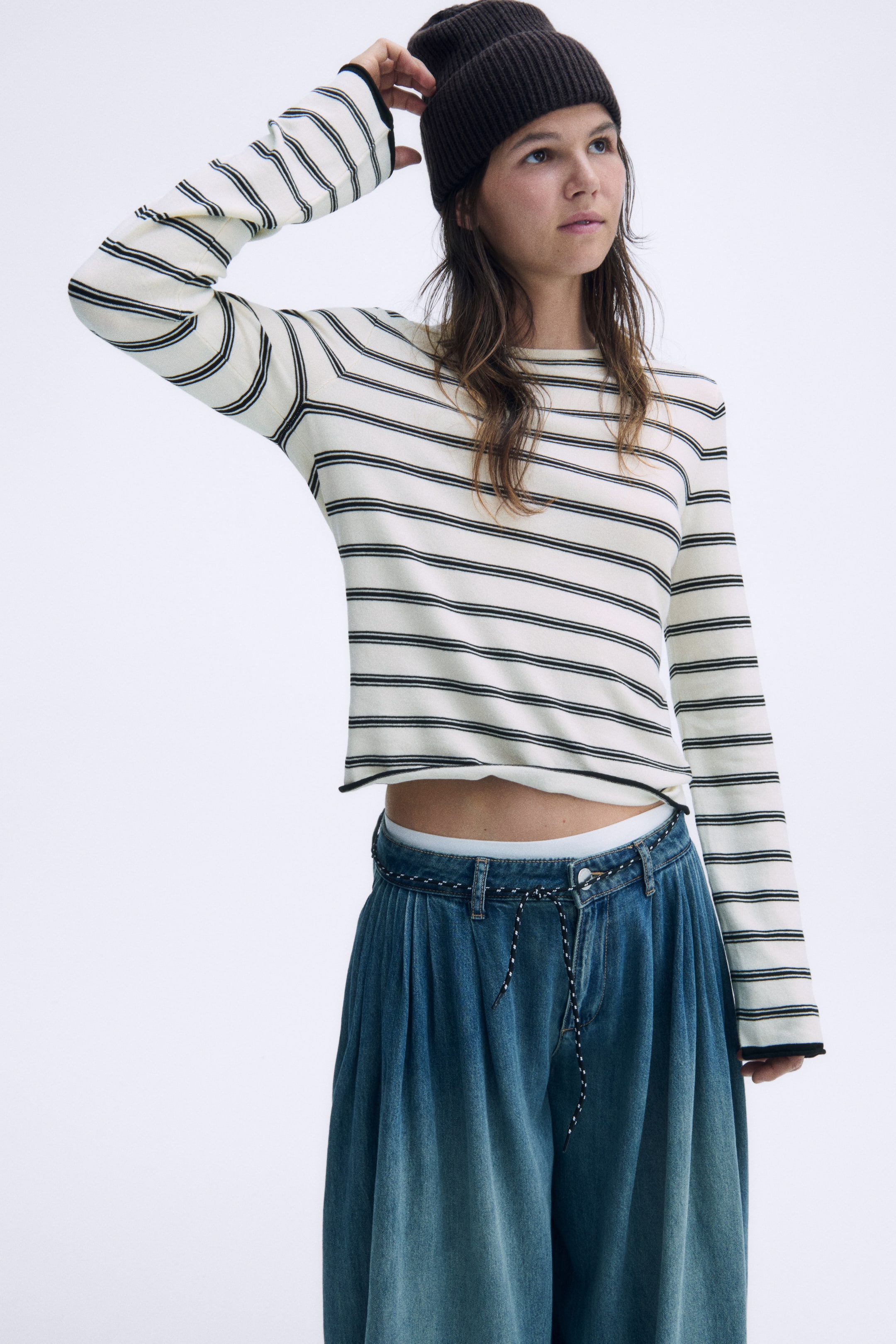 View larger image: A cream-colored, fine-knit fitted top with horizontal black stripes is facing the front, featuring a round neckline and long sleeves. The garment highlights contrasting black rolled edges at the cuffs and hem, sitting just above the waist.