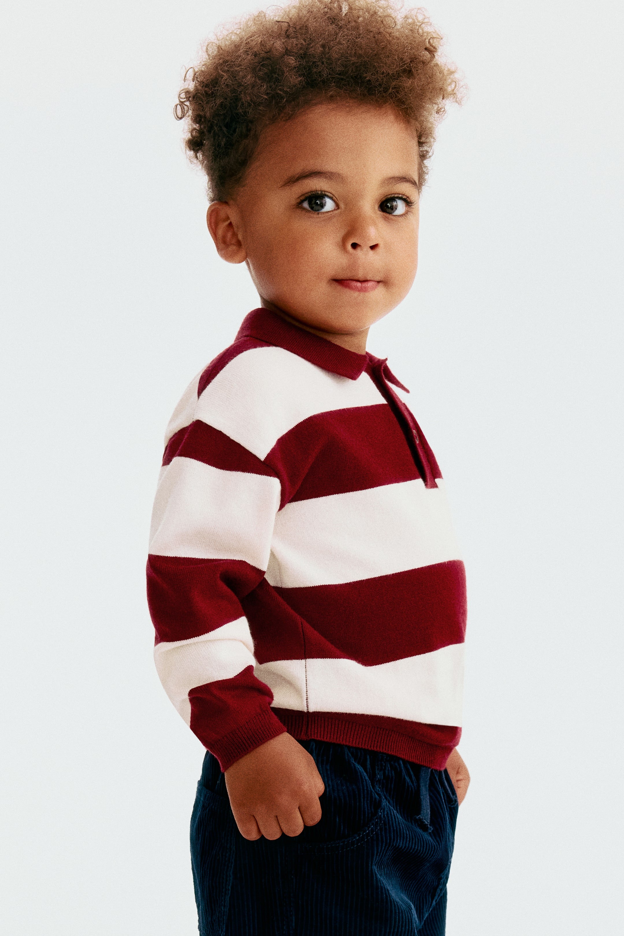 Visualizza immagine più grande: Una maglia da rugby a maniche lunghe in maglia fine, caratterizzata da ampie righe orizzontali bordeaux e bianco sporco, è mostrata da un angolo frontale a tre quarti. La maglia mette in risalto un colletto bordeaux tinta unita con abbottonatura, spalle scese e polsini e orlo a coste, abbinata a pantaloni in velluto a coste blu scuro.