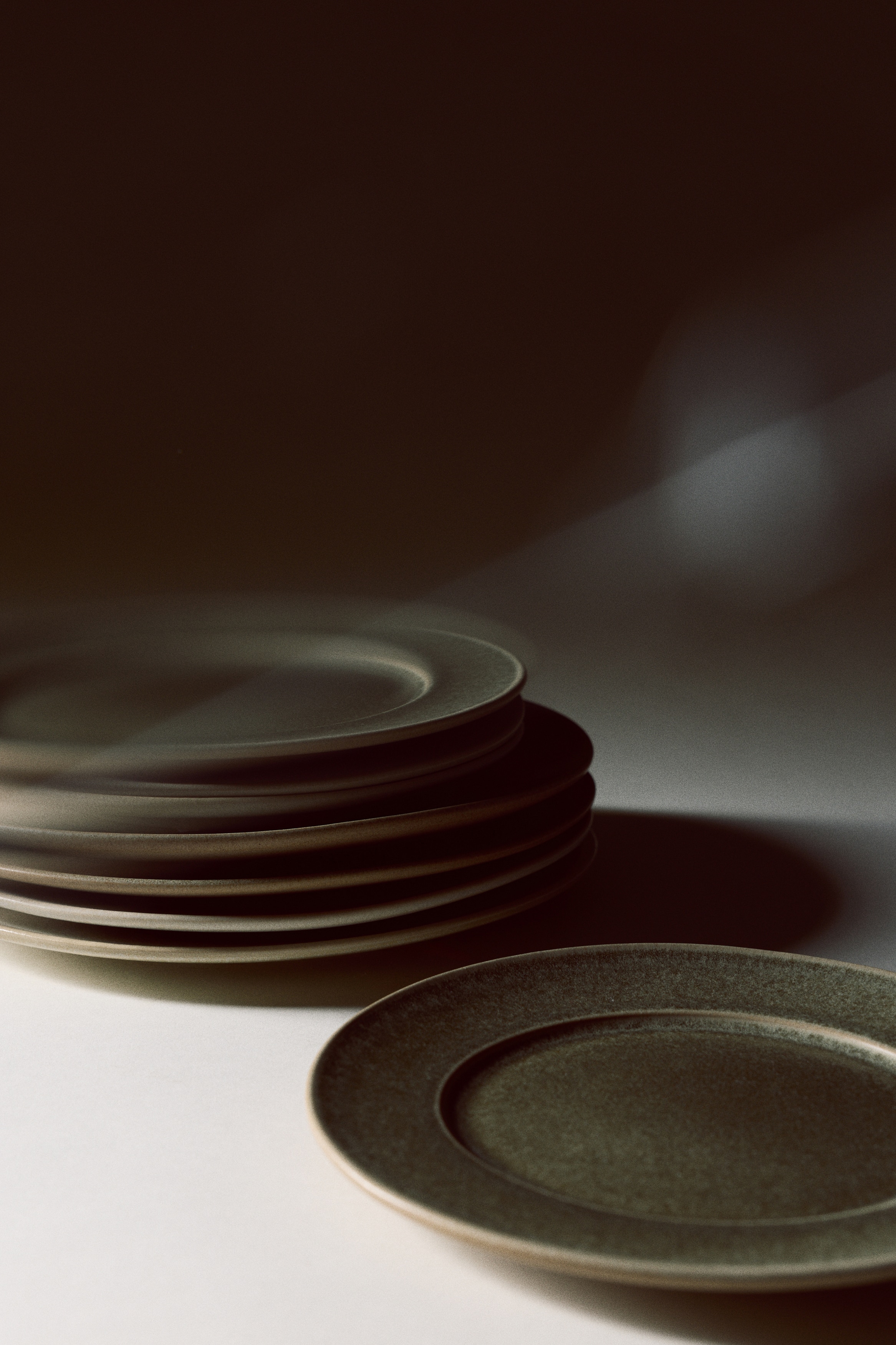 Deux assiettes rondes en cÃ©ramique sont prÃ©sentÃ©es en vue de dessus, mettant en valeur un Ã©maillage vert olive texturÃ© et mouchetÃ©. Chaque assiette prÃ©sente un creux central circulaire bien dÃ©fini ainsi quâun bord extÃ©rieur naturel de couleur brun clair.