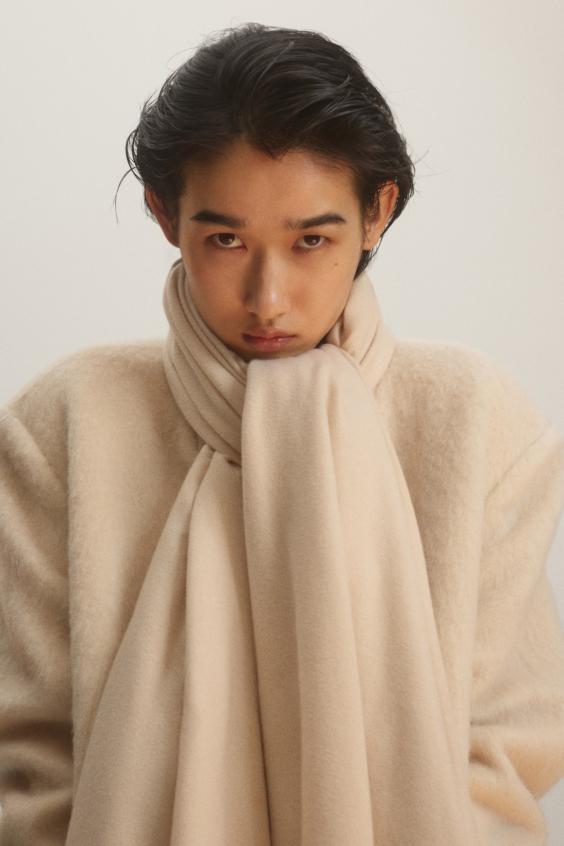 View larger image: A beige blanket scarf in a soft weave is wrapped closely around the neck, with the long ends draping down the front. The model is wearing the scarf layered over a matching cream-coloured coat.