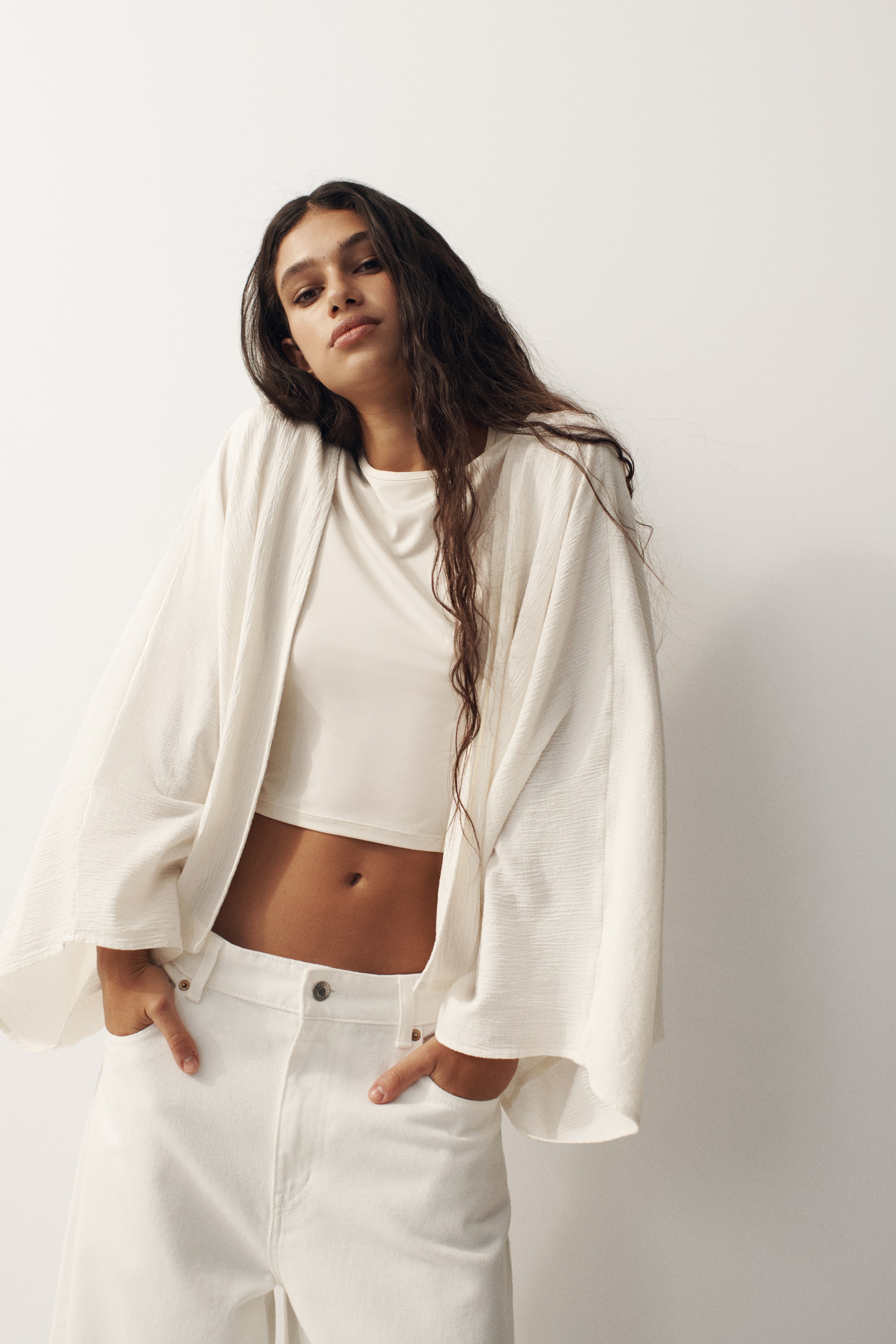 View larger image: A white open-front cardigan showcases a crinkled texture with wide, flowing three-quarter length sleeves, draped loosely over a matching crop top.