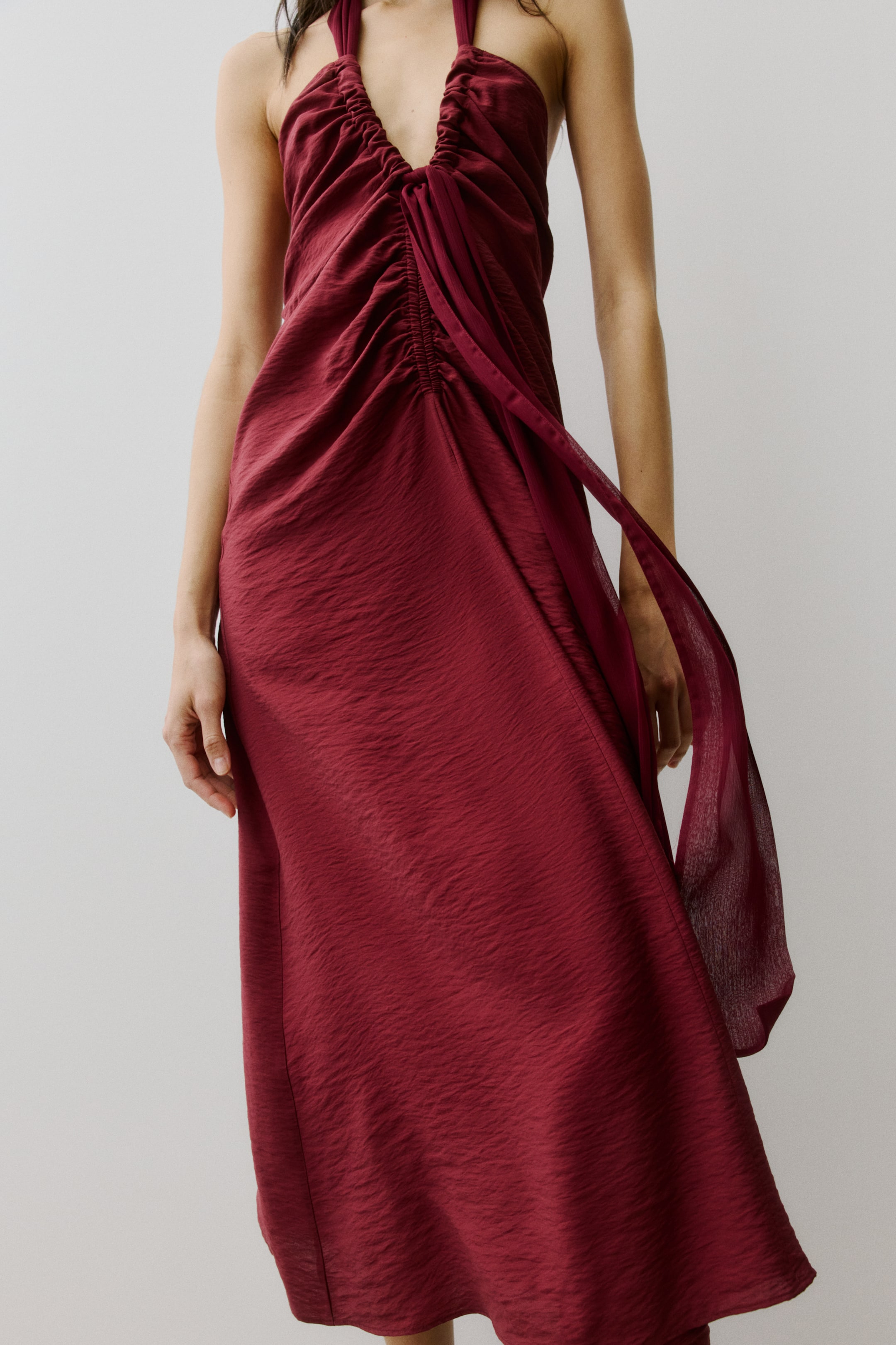 Agrandir l'image: Une robe rouge profond à col halter présente un ruché extensif sur le devant du corsage, formant un décolleté en V profond. Le tissu texturé descend gracieusement, avec un panneau drapé traversant le devant et s'étendant librement sur le côté.