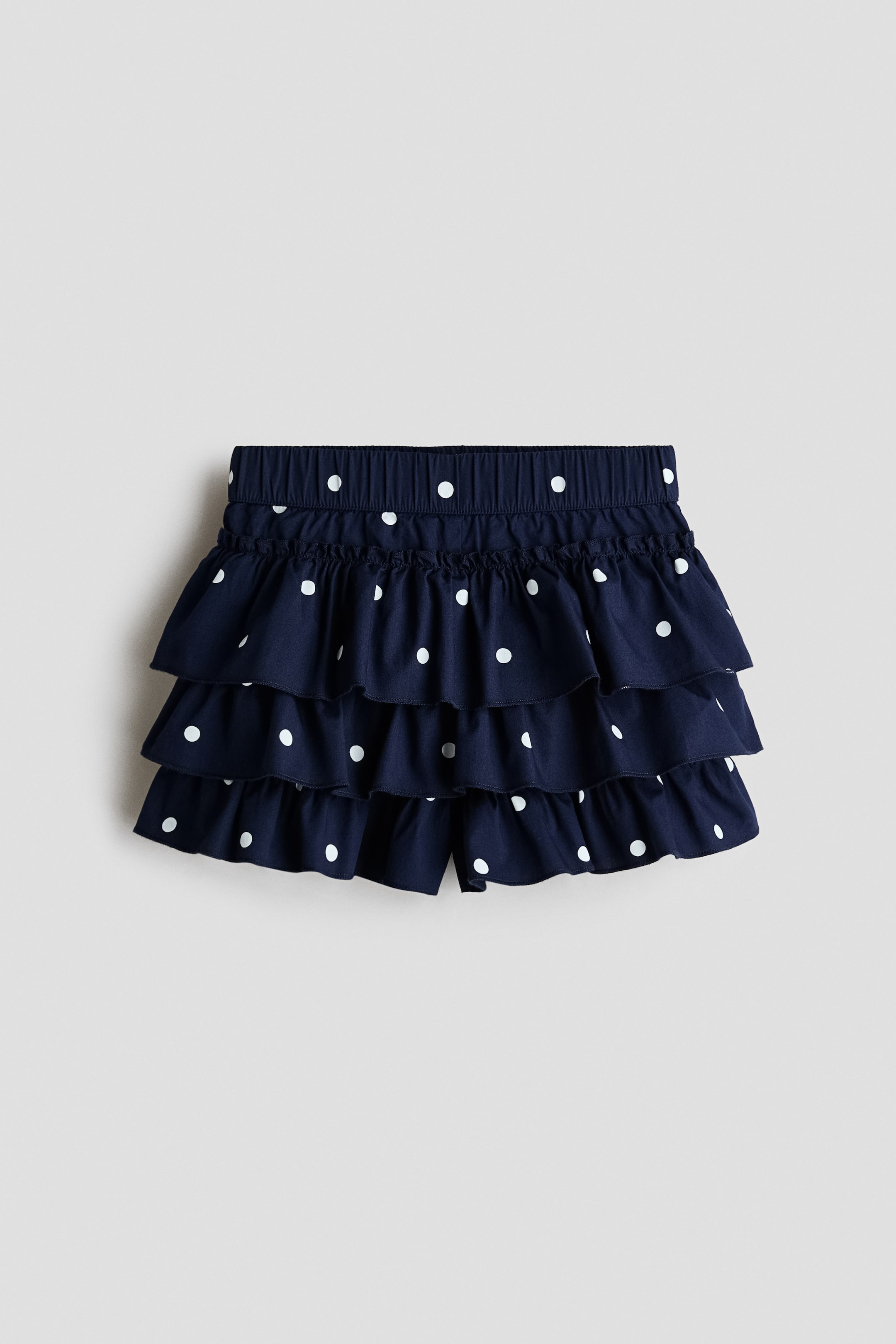 View larger image: A navy blue tiered skort with a white polka dot pattern is shown from the front, featuring an elasticated waistband with a frill trim at the top. Three tiered, ruffled layers cascade down to cover the built-in shorts.