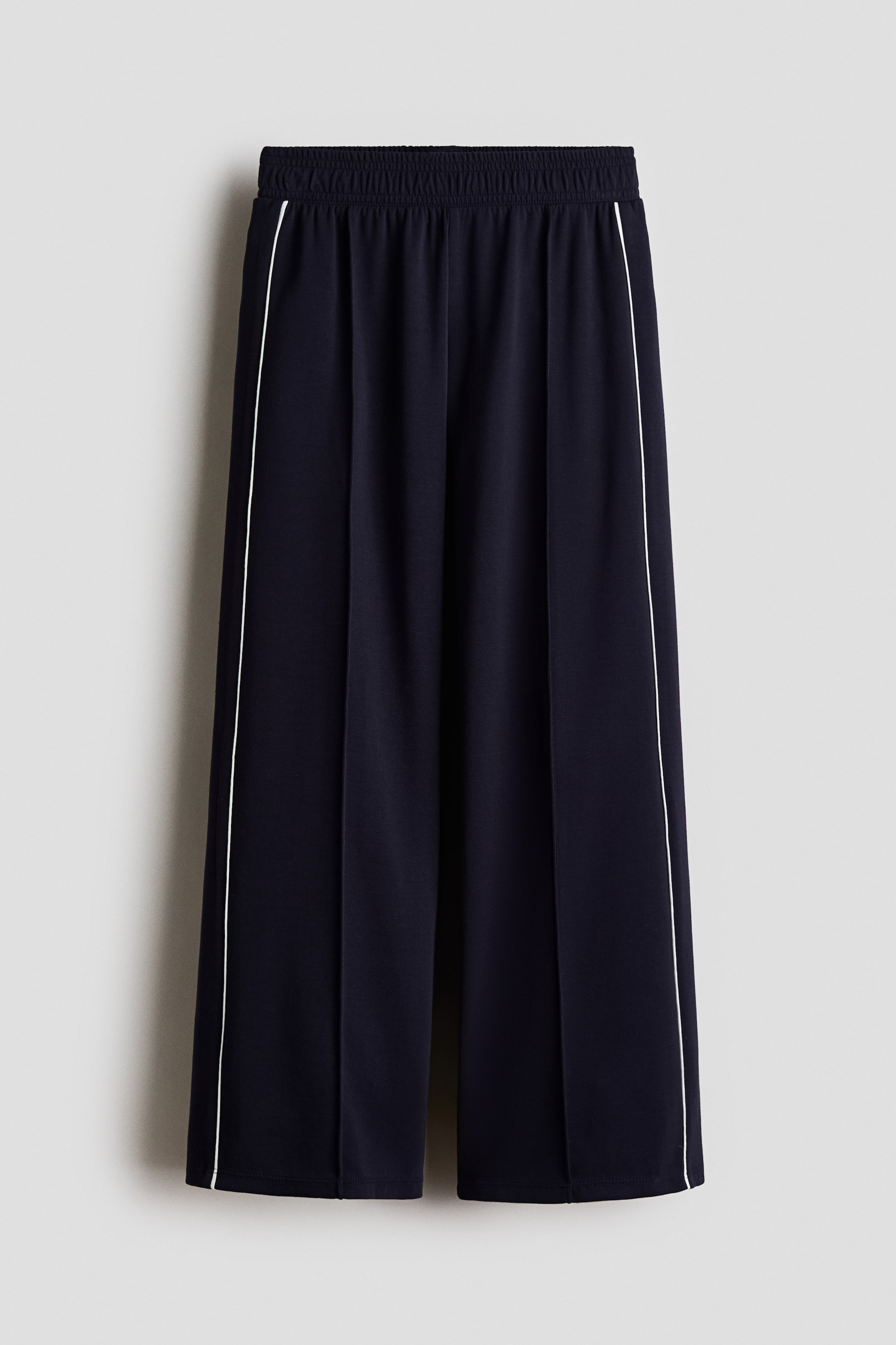 View larger image: Navy track trousers in scuba fabric are showcased from the front, featuring an elasticated waist and wide legs. Contrasting white piping runs down the sides, and sewn-in creases are visible along the front of each leg.