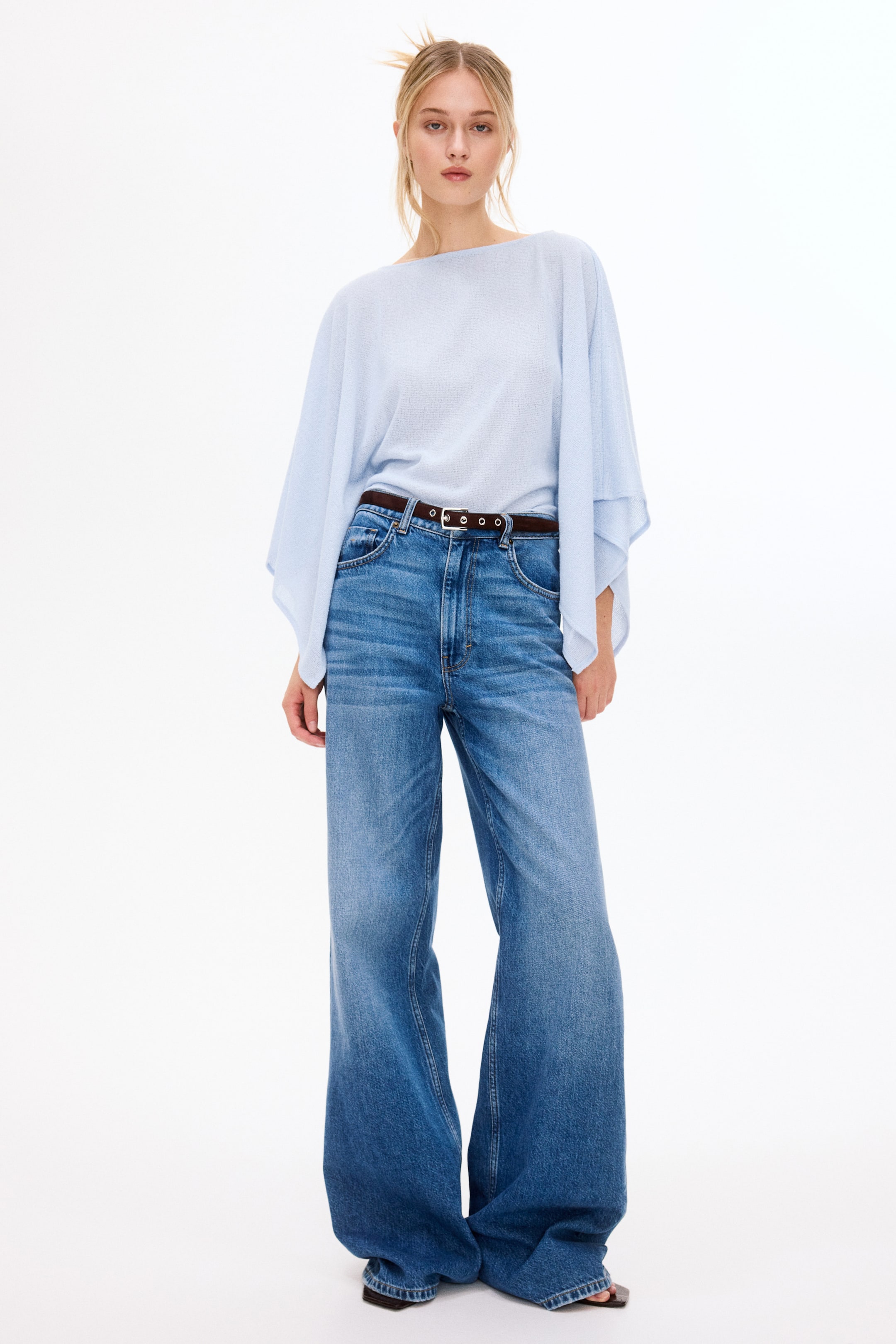 View larger image: A light blue, fine-knit boat-neck top with wide sleeves is worn, tucked into high-waisted, wide-leg blue denim jeans. A dark brown belt with silver eyelets cinches the waist.