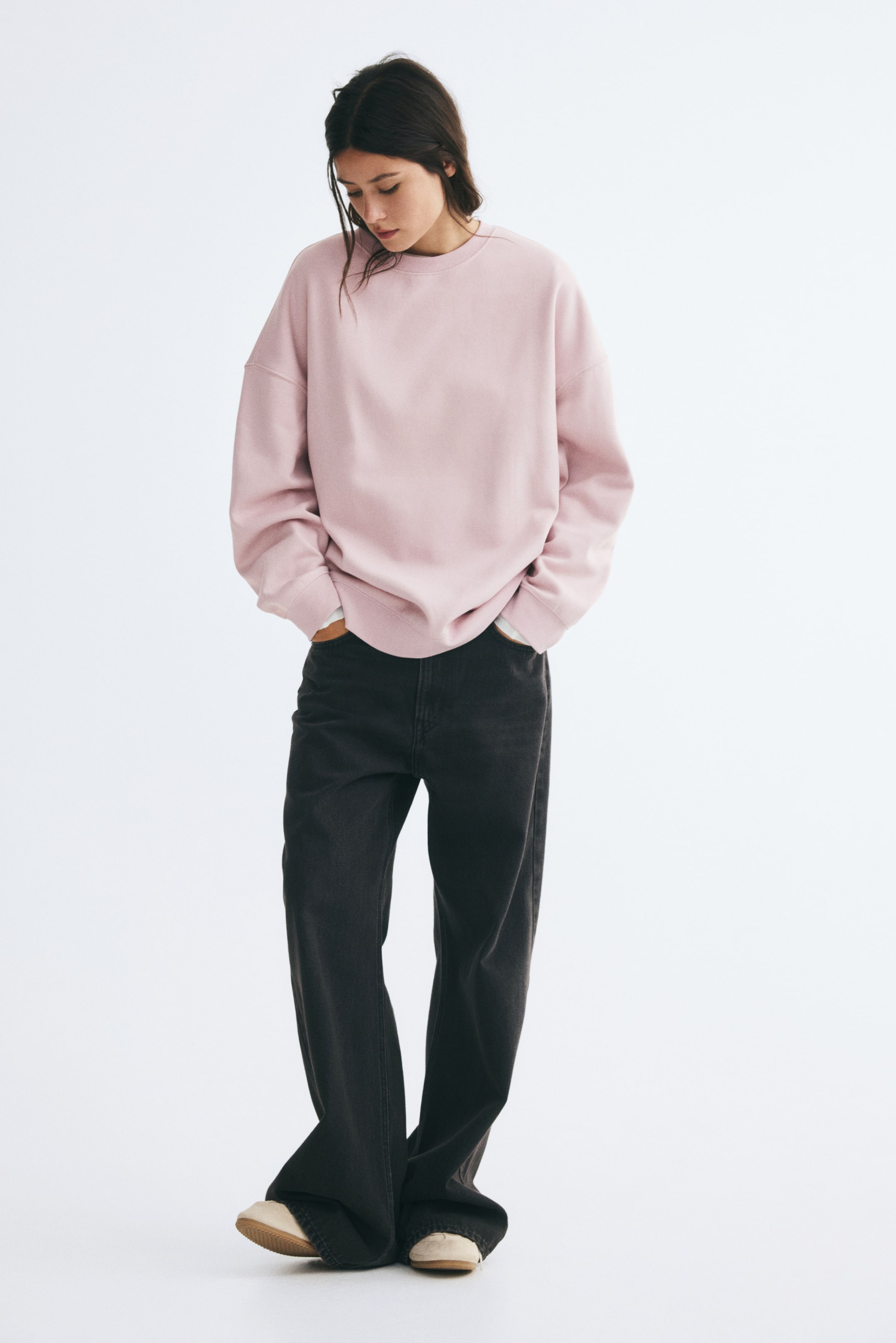 Ver imagen más grande: La modelo lleva una sudadera rosa claro de corte oversize con cuello redondo, hombros caídos y acabados de canalé. La sudadera se combina con unos vaqueros oscuros de pierna ancha, y la modelo aparece de pie con las manos metidas en los bolsillos.