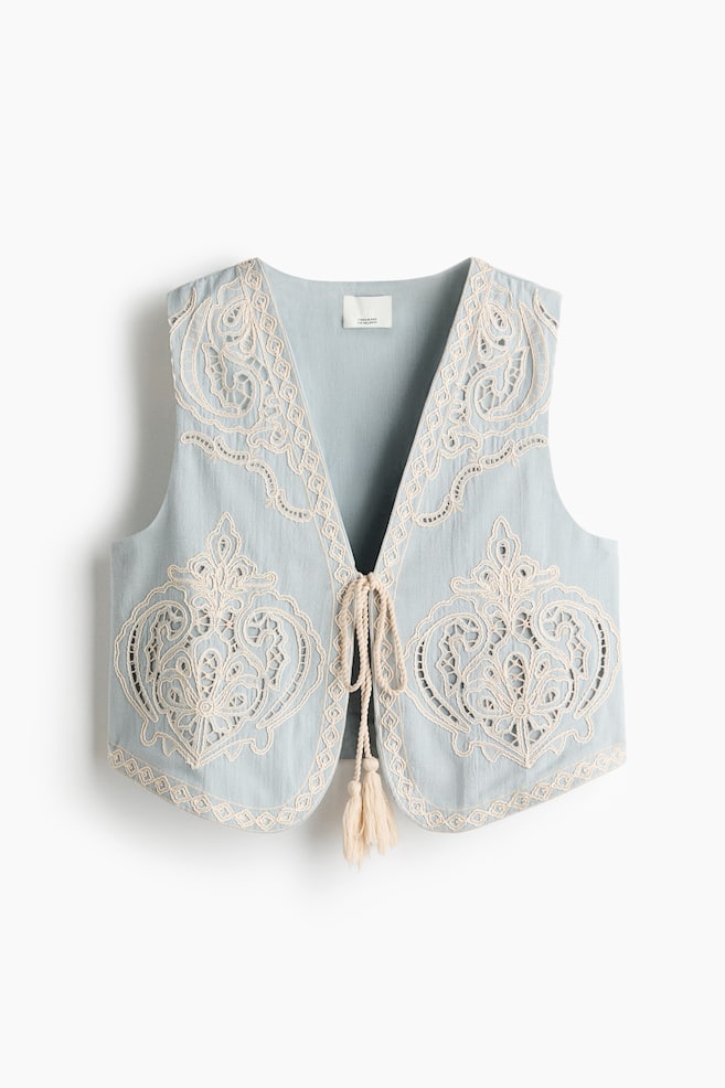 A light blue waistcoat is displayed flat, facing forward, featuring intricate cream-coloured embroidery and cutwork across its front panels, V-neckline, and armholes. It is presenting a tie-front closure with braided ties and tassels. - 1