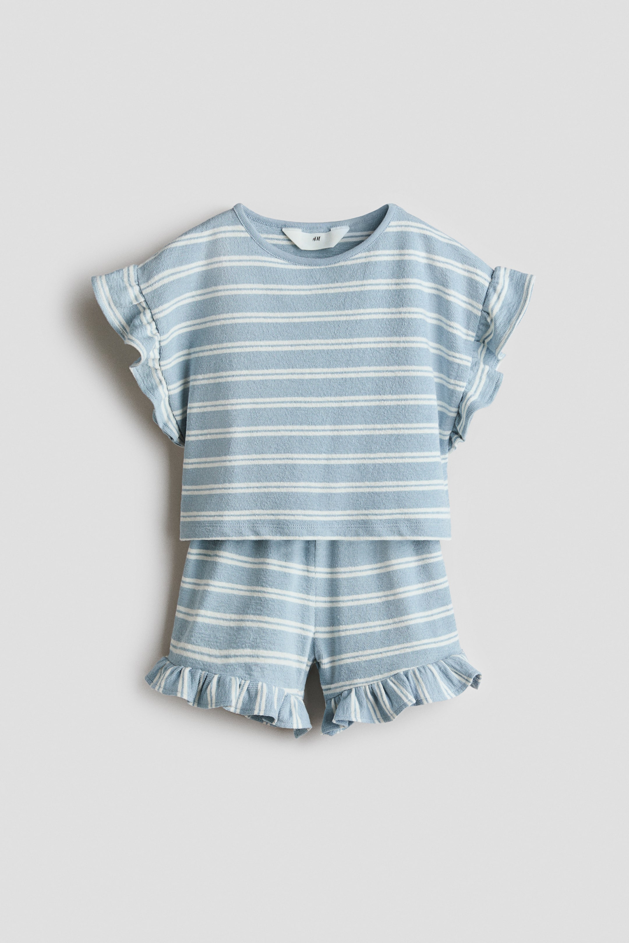 拡大画像を表示: ライトブルーとホワイトのストライプ柄のテリーセットは、丸いトリム付きネックライン、ドロップショルダー、フリル付きのショートスリーブが特徴です。マッチングショーツは、ゴム入りのドローストリングウエストとフリル付きの裾が施されています。