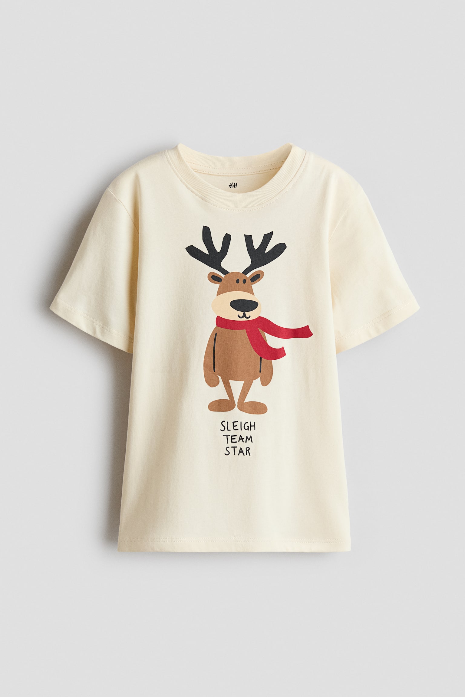 A cream-coloured, short-sleeved T-shirt is shown from the front, featuring a ribbed crew neck and a central graphic of a cartoon reindeer. The reindeer is wearing a red scarf, and the text "SLEIGH TEAM STAR" appears in black print directly below the illustration.