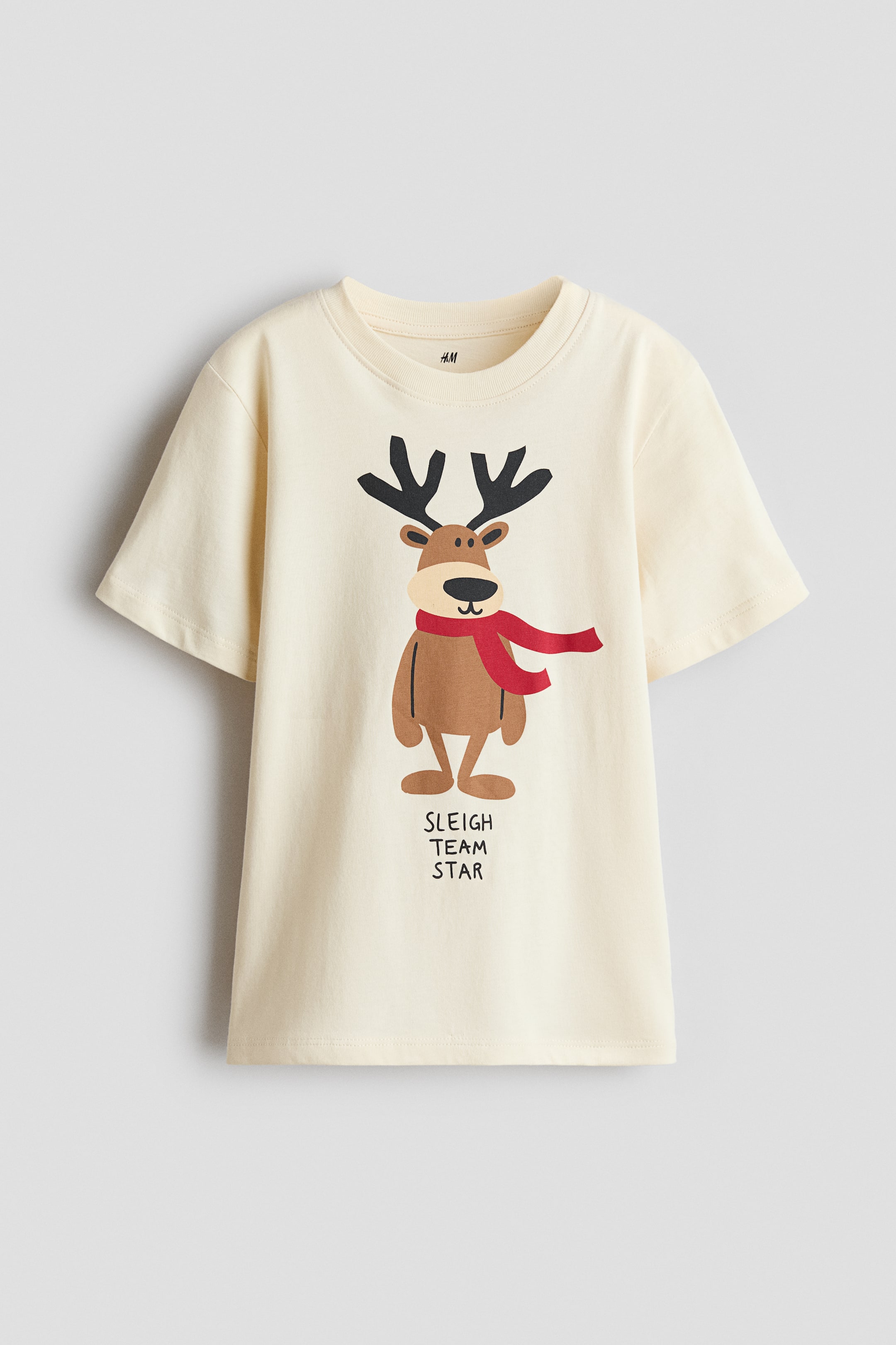 View larger image: A cream-coloured, short-sleeved T-shirt is shown from the front, featuring a ribbed crew neck and a central graphic of a cartoon reindeer. The reindeer is wearing a red scarf, and the text "SLEIGH TEAM STAR" appears in black print directly below the illustration.