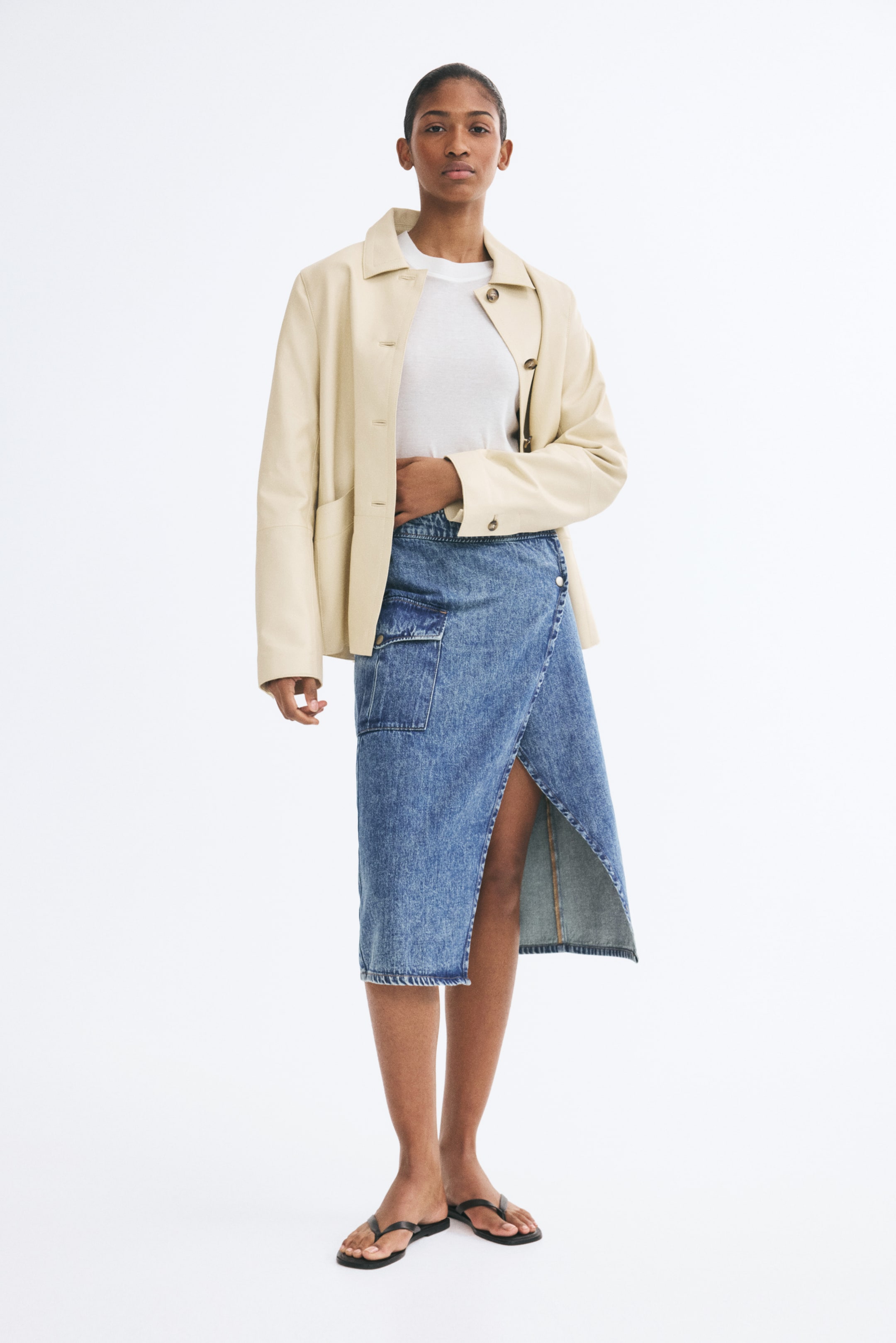 View larger image: The model is wearing a calf-length denim wrap skirt featuring an asymmetrical front overlap and a bellows pocket with a flap on one side. The skirt is styled with a white T-shirt, a cream-colored jacket, and black thong sandals.
