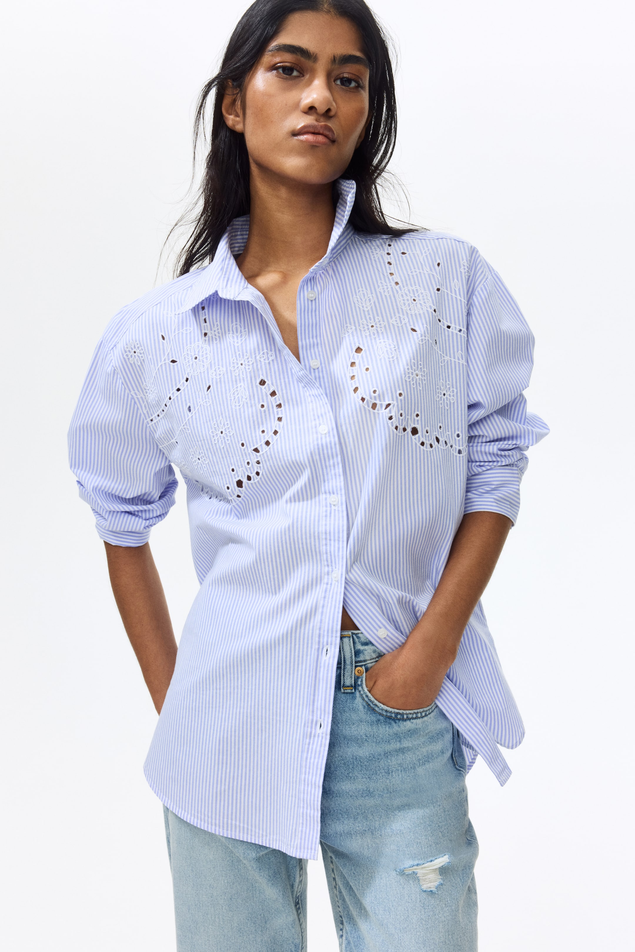 View larger image: A blue and white striped shirt is showcased in a front-angled view, featuring intricate white eyelet embroidery with floral and curved motifs across the chest. It is designed with a standing pointed collar and long sleeves rolled up for a casual look. The front placket is partially open, adding a relaxed vibe. One side of the hem is styled tucked into light-wash jeans, while the other side is left hanging loose, offering a chic and effortless appearance.