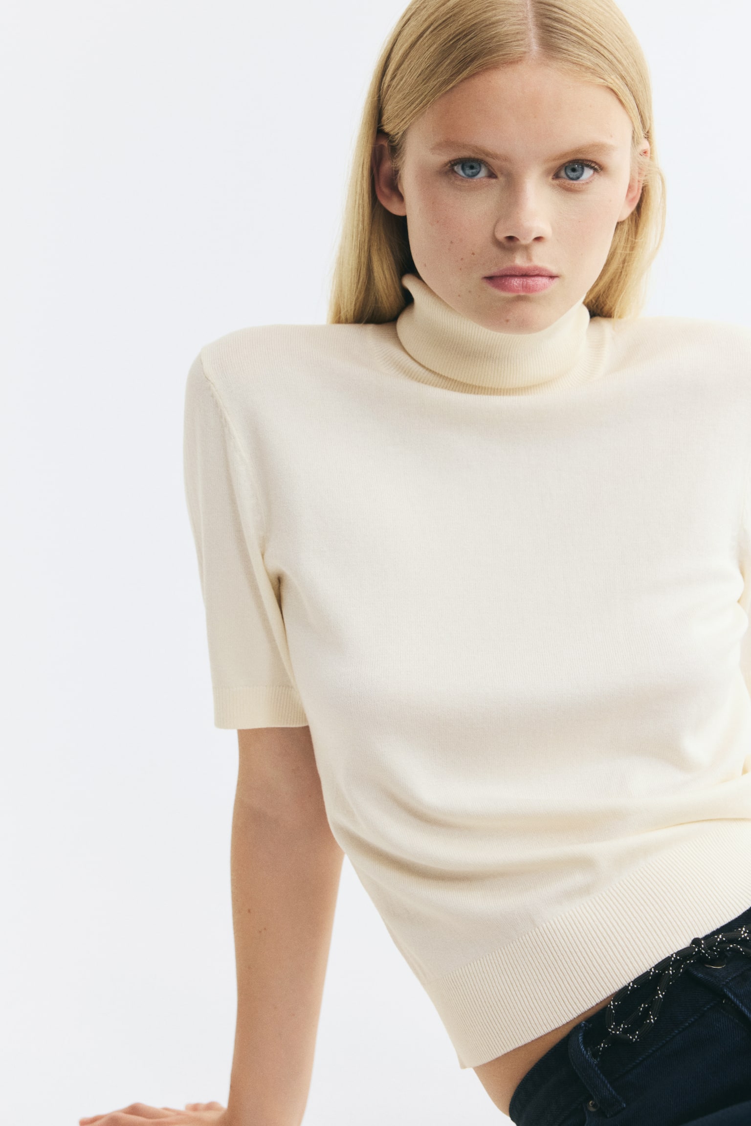 The model is wearing a cream-coloured fine-knit top featuring a ribbed turtleneck, short sleeves with ribbed cuffs, and a ribbed hem. The top is paired with dark denim trousers visible at the waist. - 1