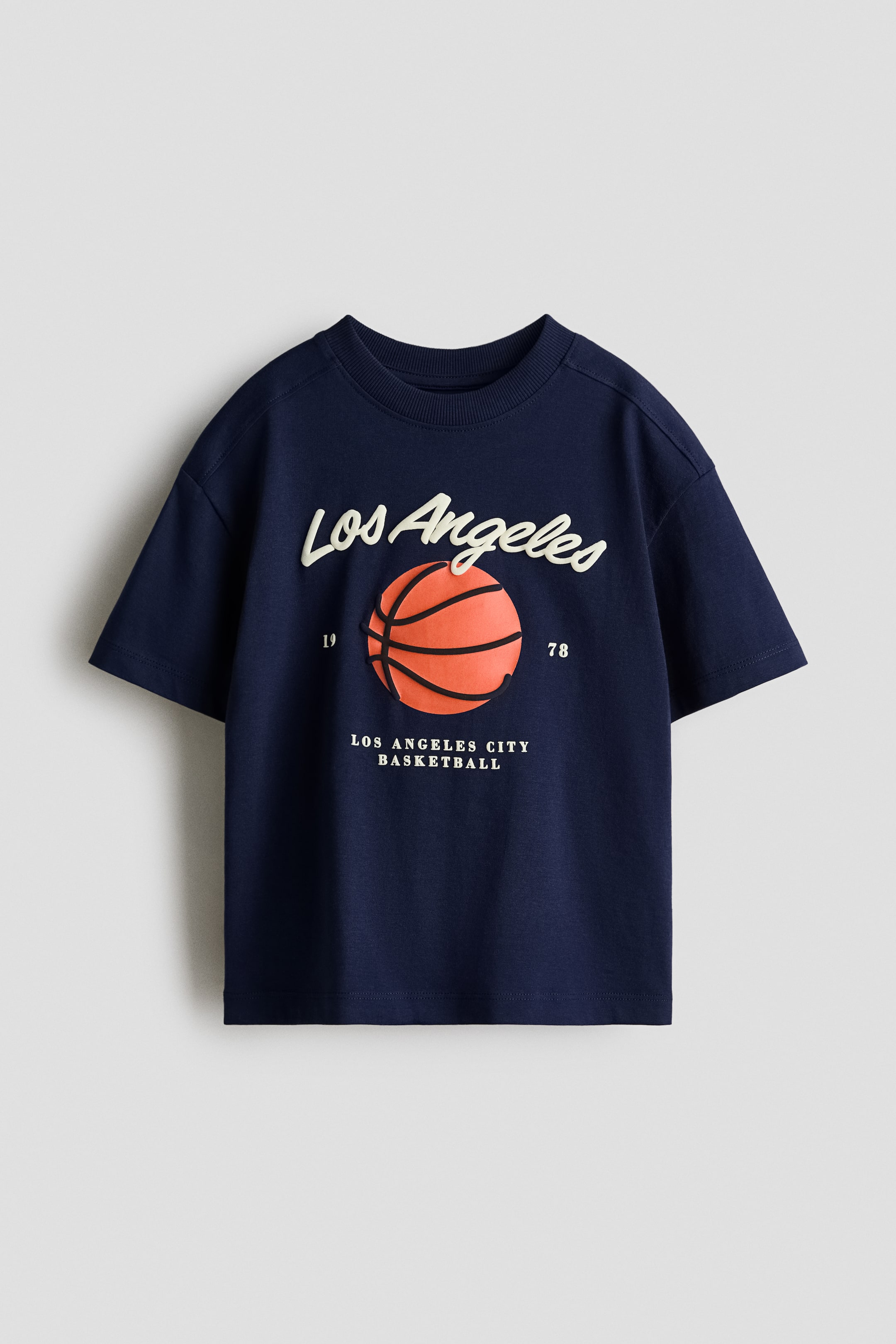 View larger image: A navy blue t-shirt is facing front, featuring a ribbed crew neck and short sleeves. It is displaying a graphic print with "Los Angeles" in white script, an orange basketball, the numbers "19" and "78", and the text "LOS ANGELES CITY BASKETBALL" below.